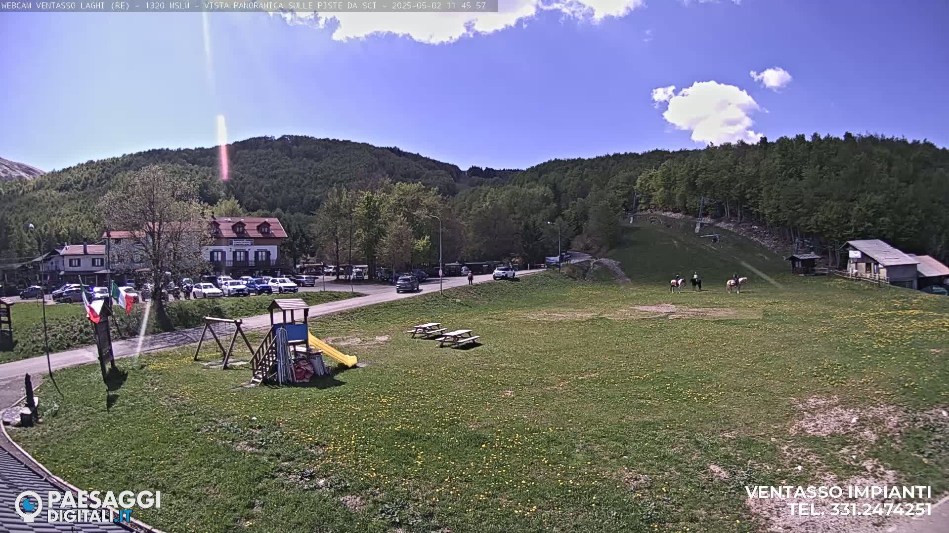 A sunny day reveals a grassy field with a playground, picnic tables, a parking lot full of cars, a building, and people riding horses, all nestled amidst a green, mountainous landscape.