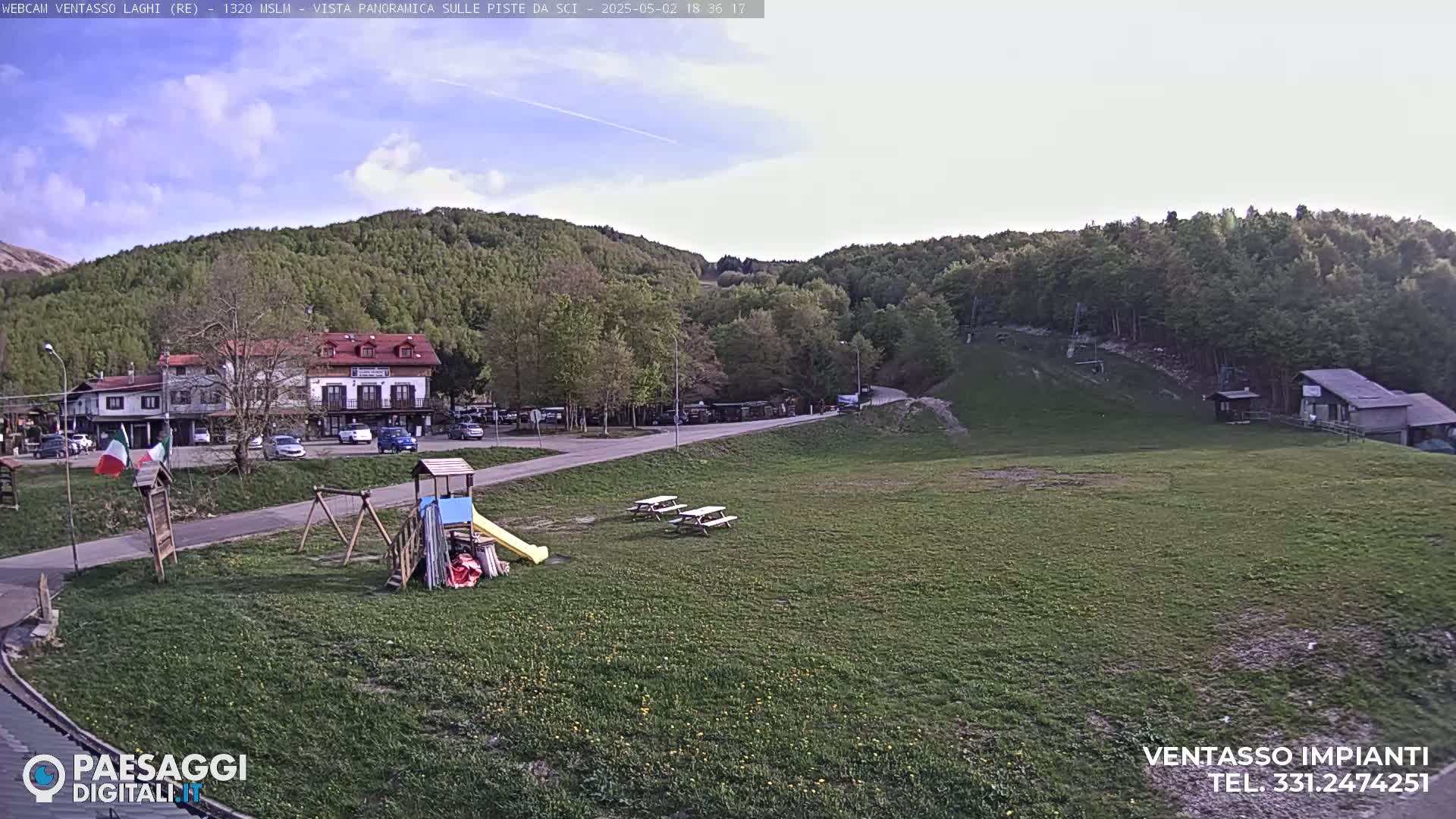 A grassy area with picnic tables and a playground sits in front of a building and parking lot, all nestled at the base of a wooded hill under a mostly clear sky.