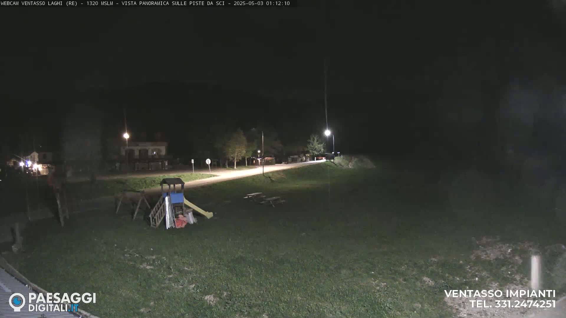A nighttime view of a grassy area with a playground and picnic tables near a road, illuminated by streetlights.
