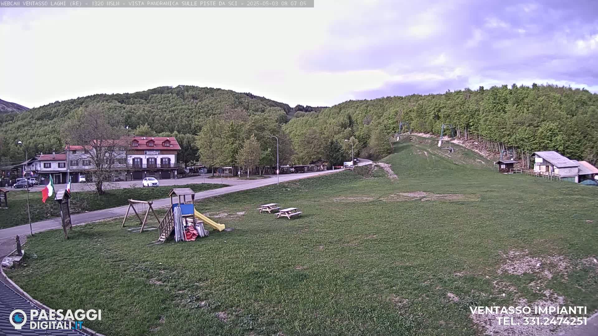 A playground and picnic tables sit in a grassy area overlooking a village nestled among green hills under a partly cloudy sky.