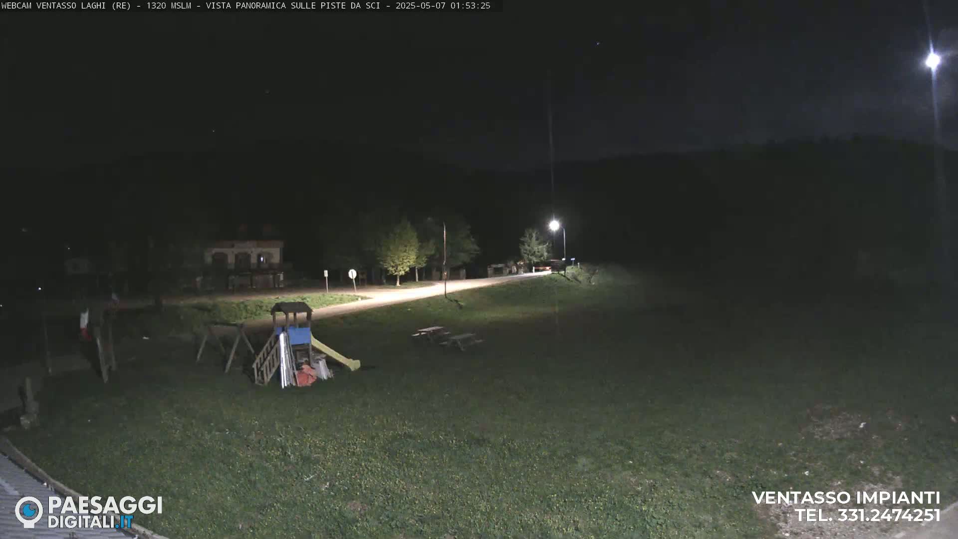A nighttime view of a grassy area with a playground and picnic tables, illuminated by a single streetlight, beside a road.