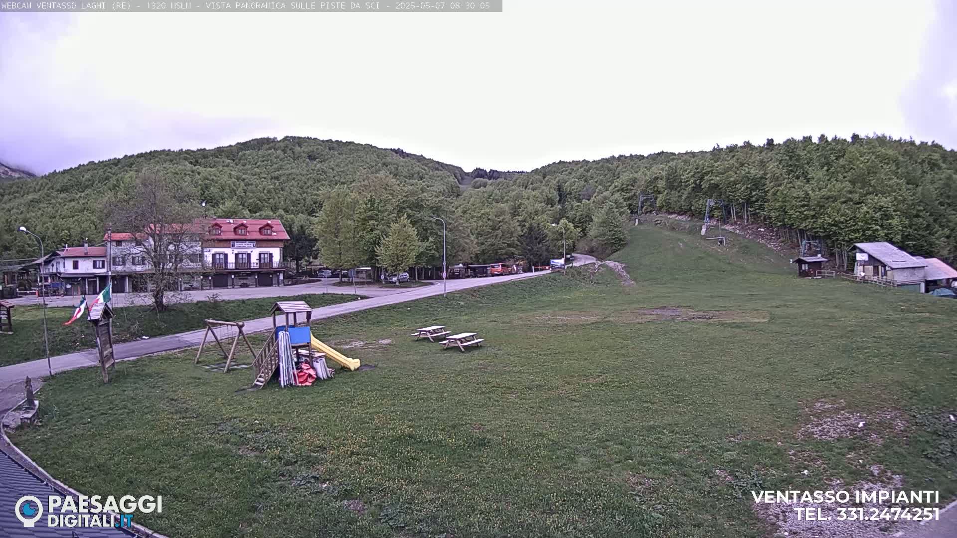 A mostly cloudy day reveals a mountainside village with buildings, a playground, picnic tables, and a ski lift, all surrounded by green hills and forests.