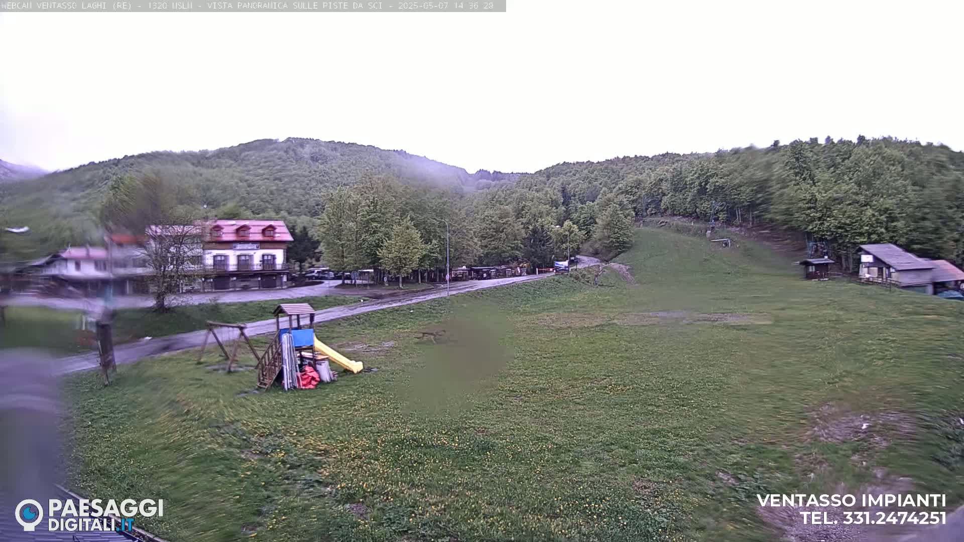 A playground sits in a grassy field overlooking a village nestled in a hillside forest under an overcast sky.
