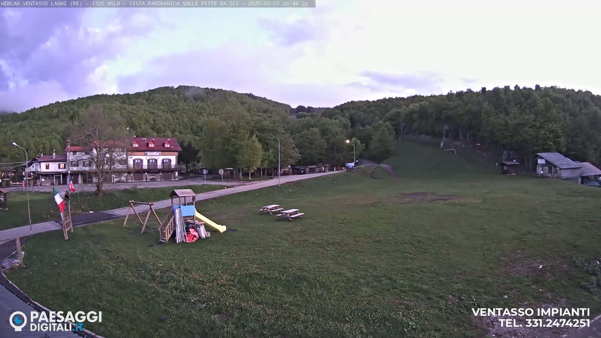 A playground and picnic tables sit in a grassy area near a building and forested hillside under a partly cloudy sky.