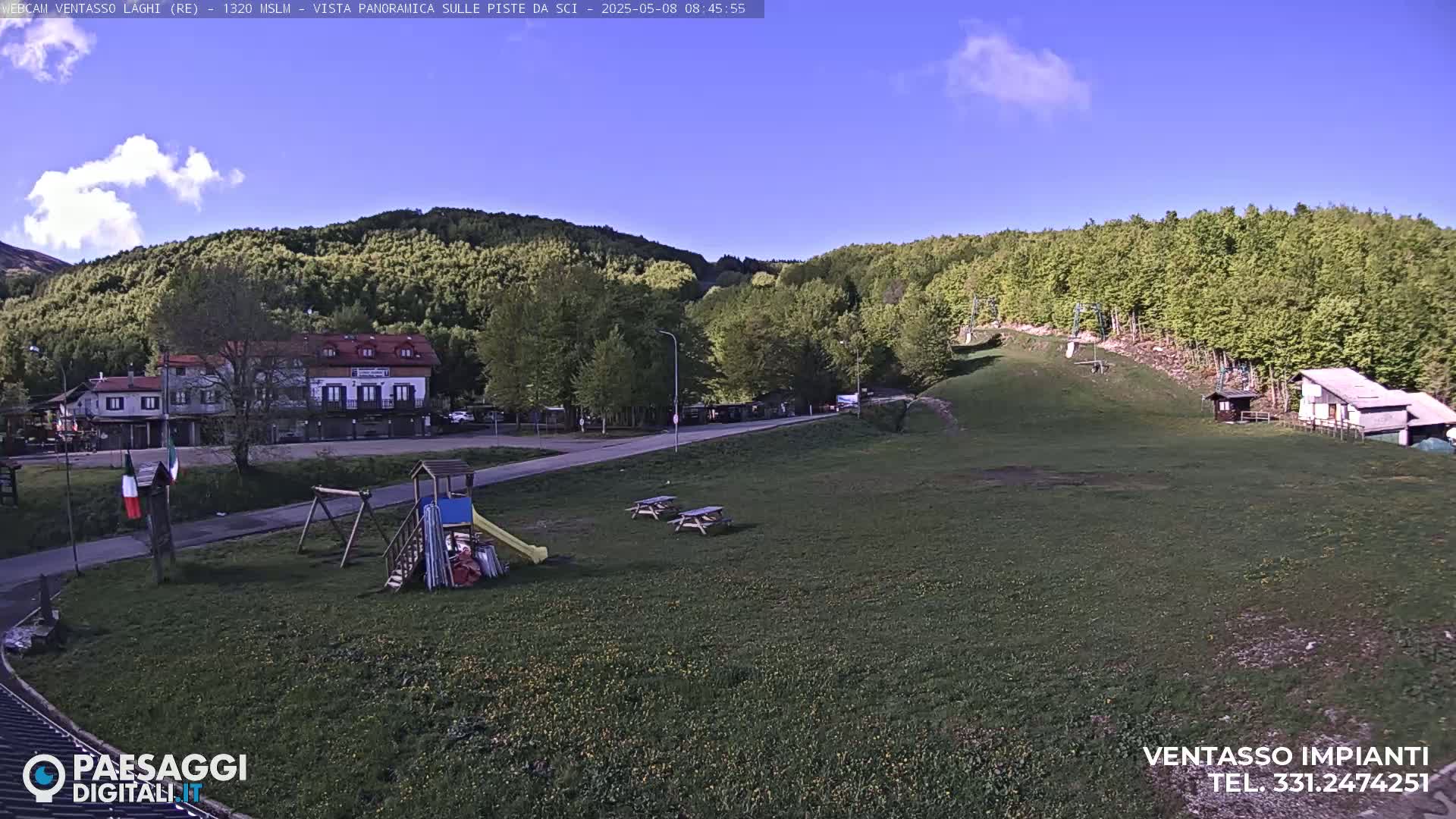 A sunny day reveals a grassy area with a playground, picnic tables, a building, and a ski lift against a backdrop of green hills and a clear blue sky.