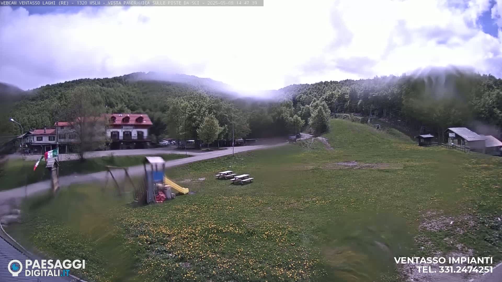 A partly cloudy mountain vista shows a grassy area with picnic tables, a playground, and a building, with fog partially obscuring the mountainside in the background.
