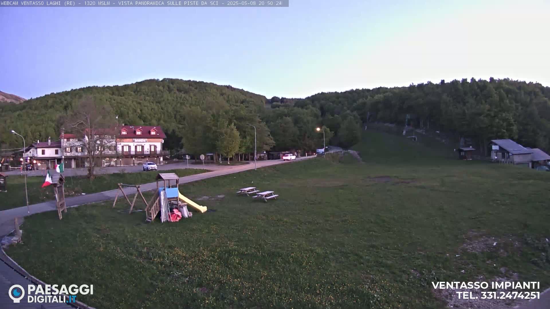 A playground and picnic tables sit in a grassy area near a building and forested hills under a twilight sky.