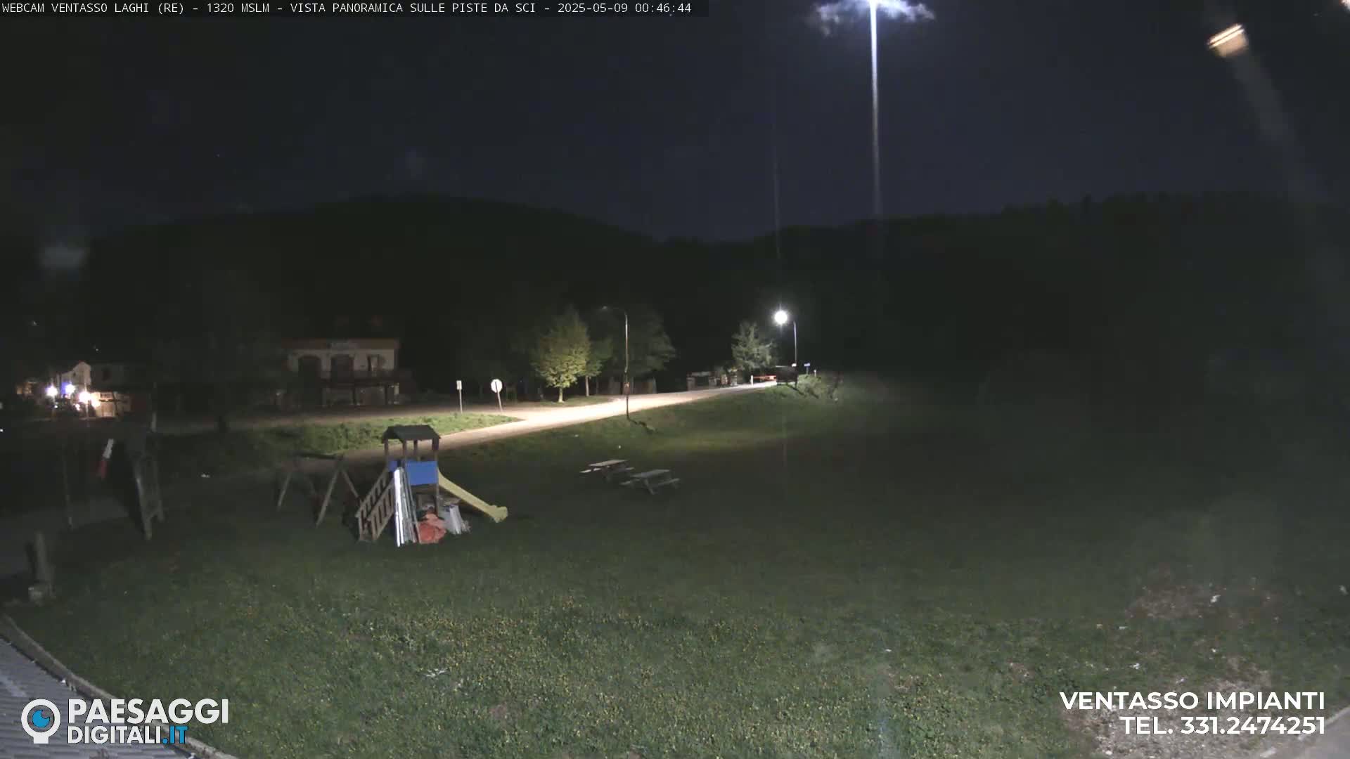 A nighttime view of a playground and picnic tables in a grassy area next to a road, under partly cloudy skies.