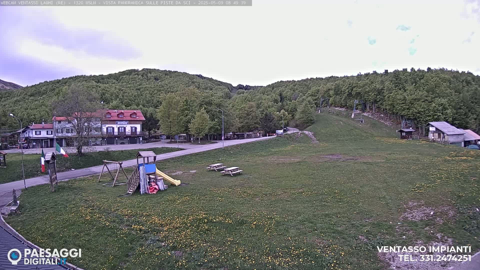 A playground and picnic tables sit in a grassy field below a building and a wooded hillside on an overcast day.