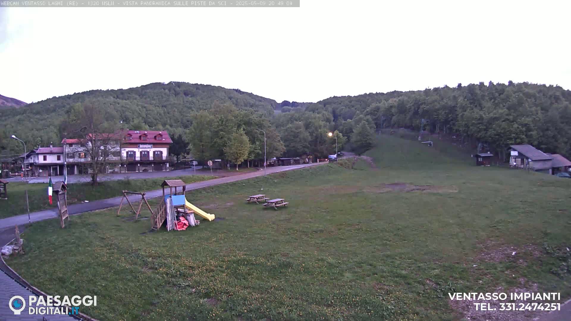 A grassy area with a playground, picnic tables, and buildings sits below a hillside of green trees under an overcast sky.