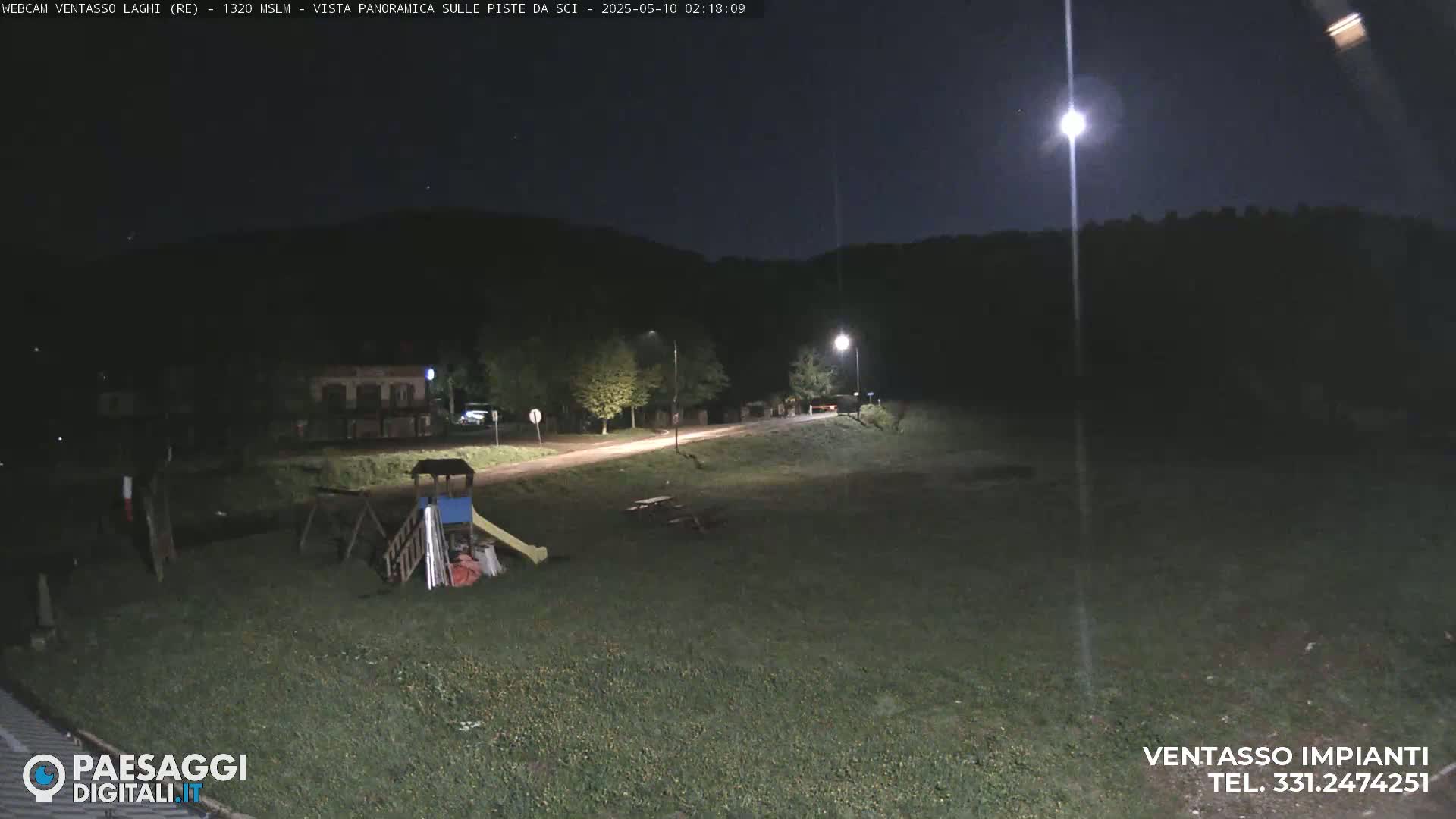 A nighttime view of a grassy area with a playground, a road, and distant trees under a bright moon.