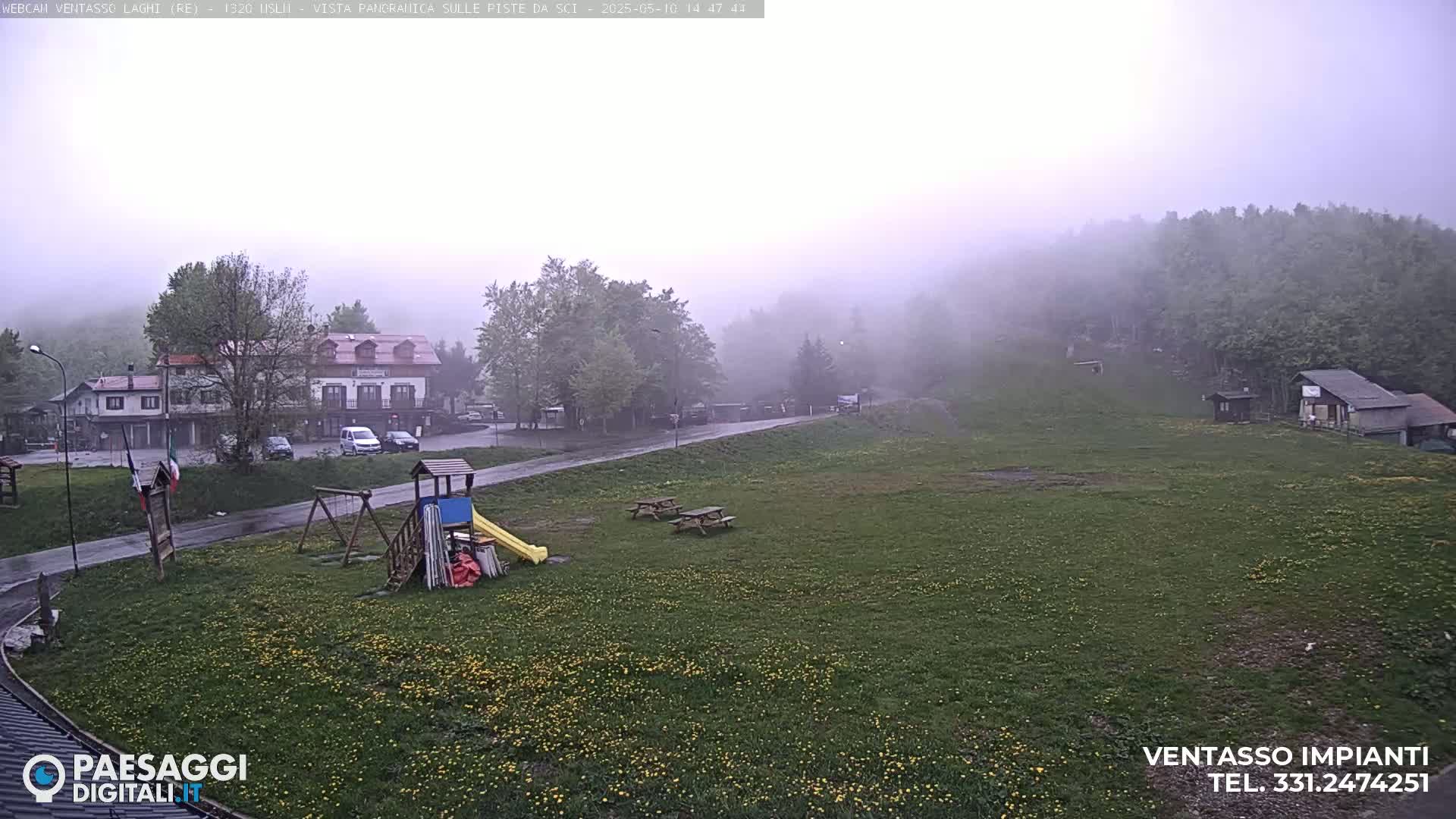 A foggy, rainy day reveals a grassy field with picnic tables and playground equipment near a small village nestled among misty hills.
