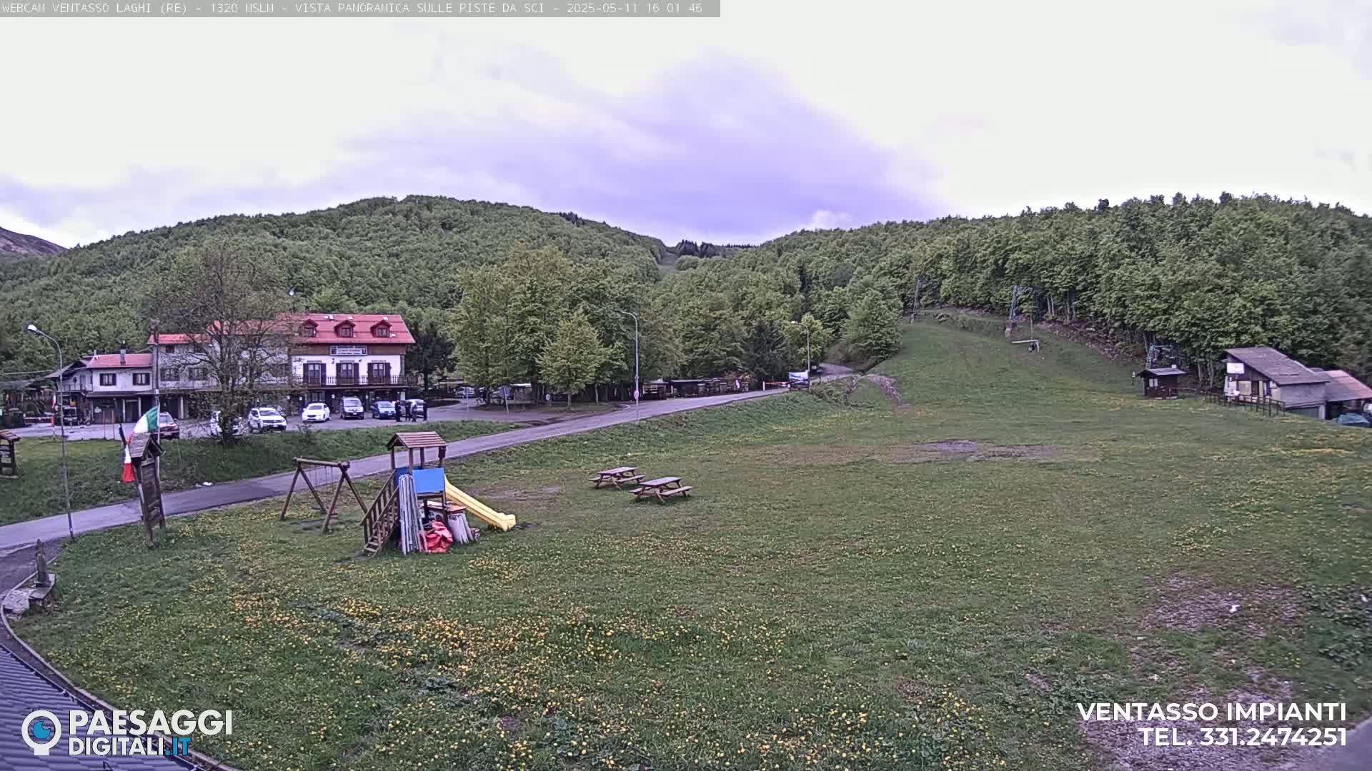 A grassy field with picnic tables, a playground, a building, and a wooded hillside under an overcast sky.