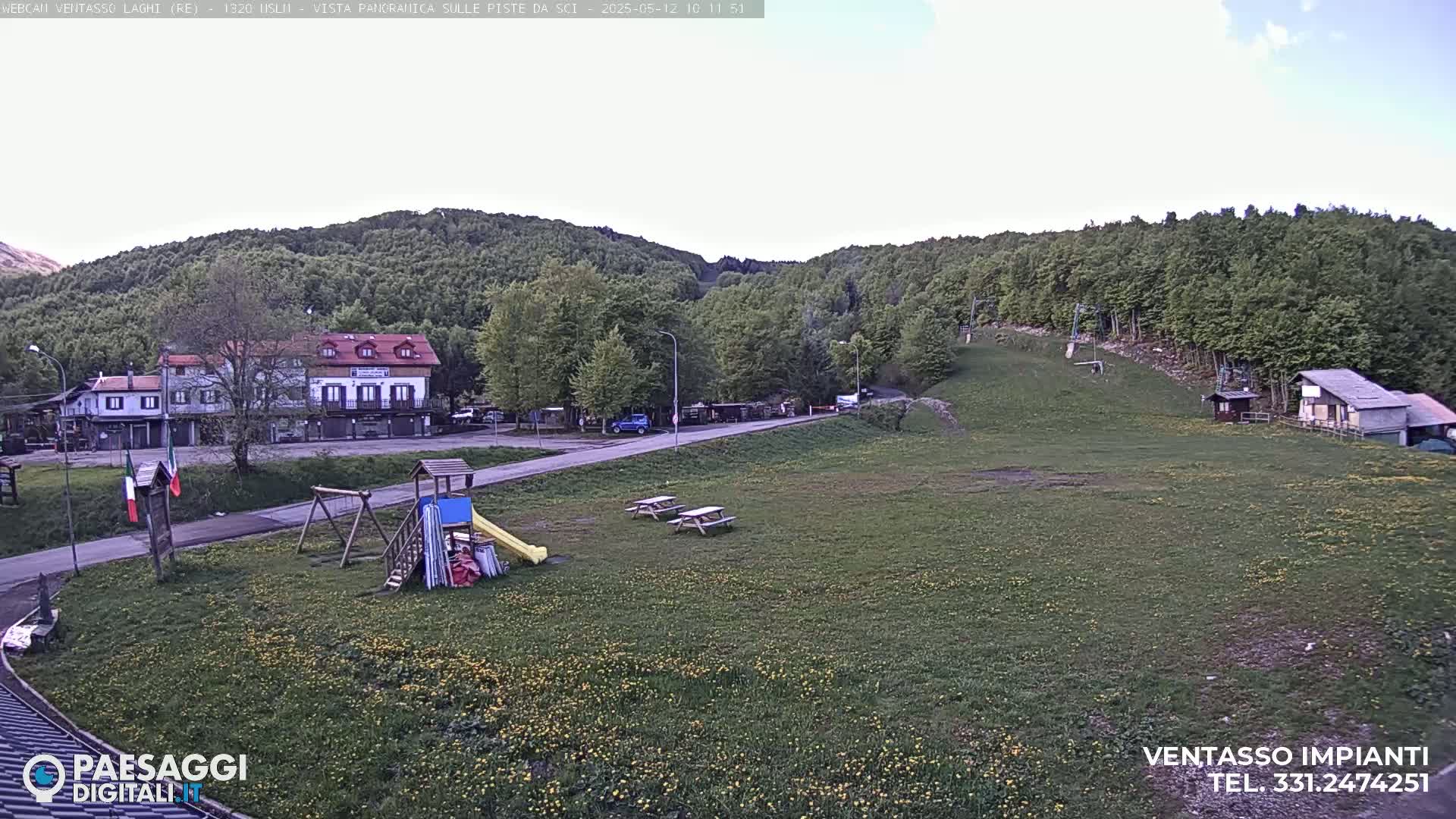 A grassy field with a playground, picnic tables, and buildings sits in front of a hillside covered in green trees under a mostly clear sky.