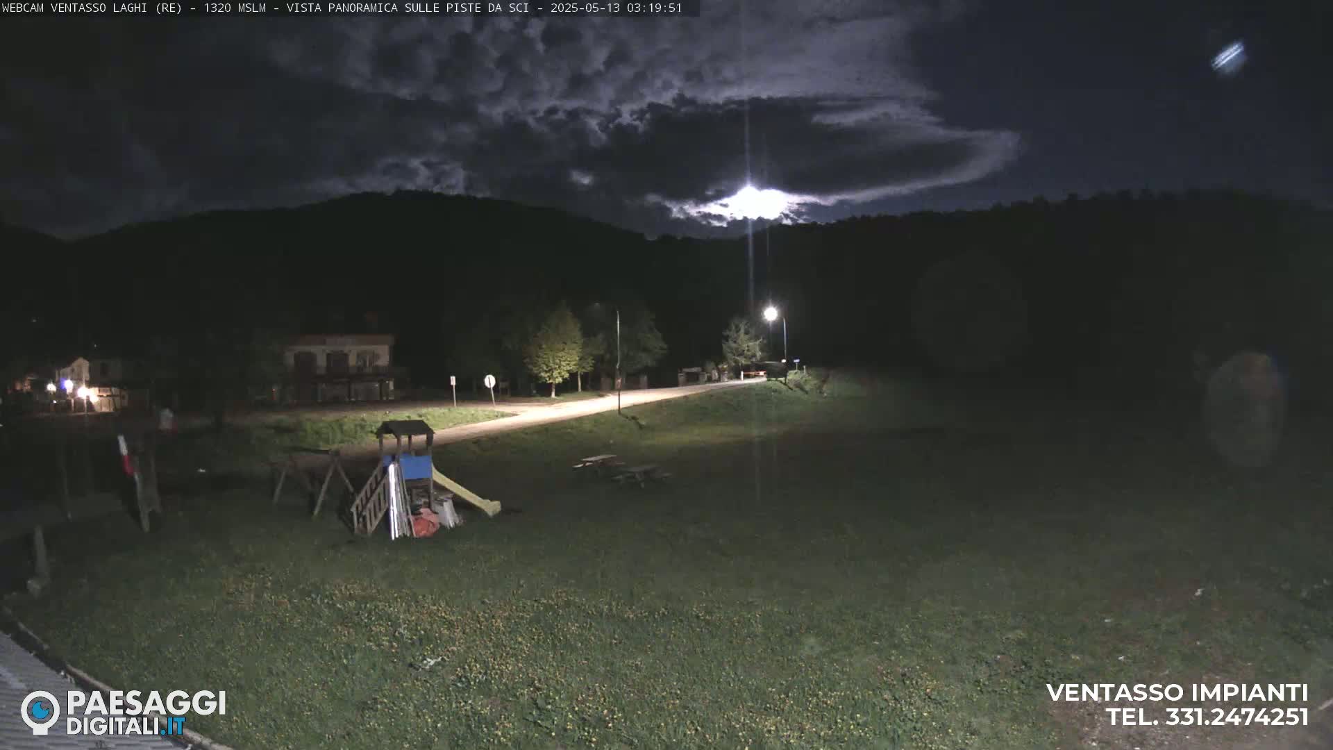 A nighttime view of a grassy area with playground equipment, a road, and buildings in the background under a partly cloudy sky.