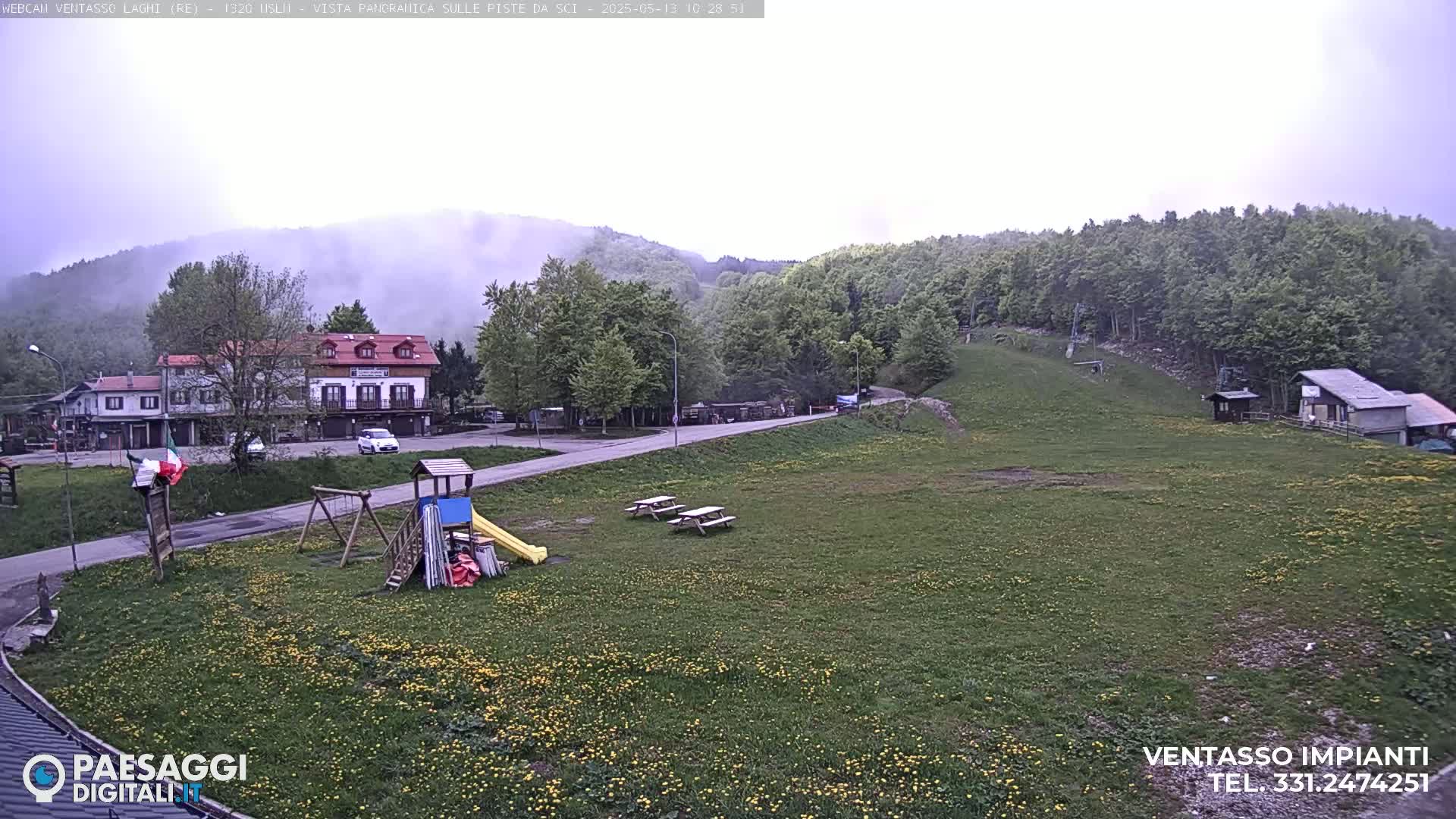A grassy field with a playground, picnic tables, and a building sits below a hillside of trees in foggy conditions.
