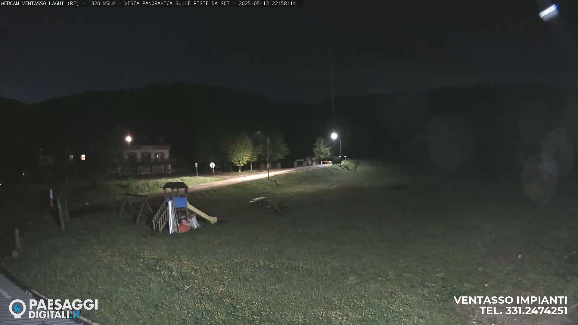 A nighttime view of a playground and grassy area near a road, illuminated by streetlights, under a clear, dark sky.