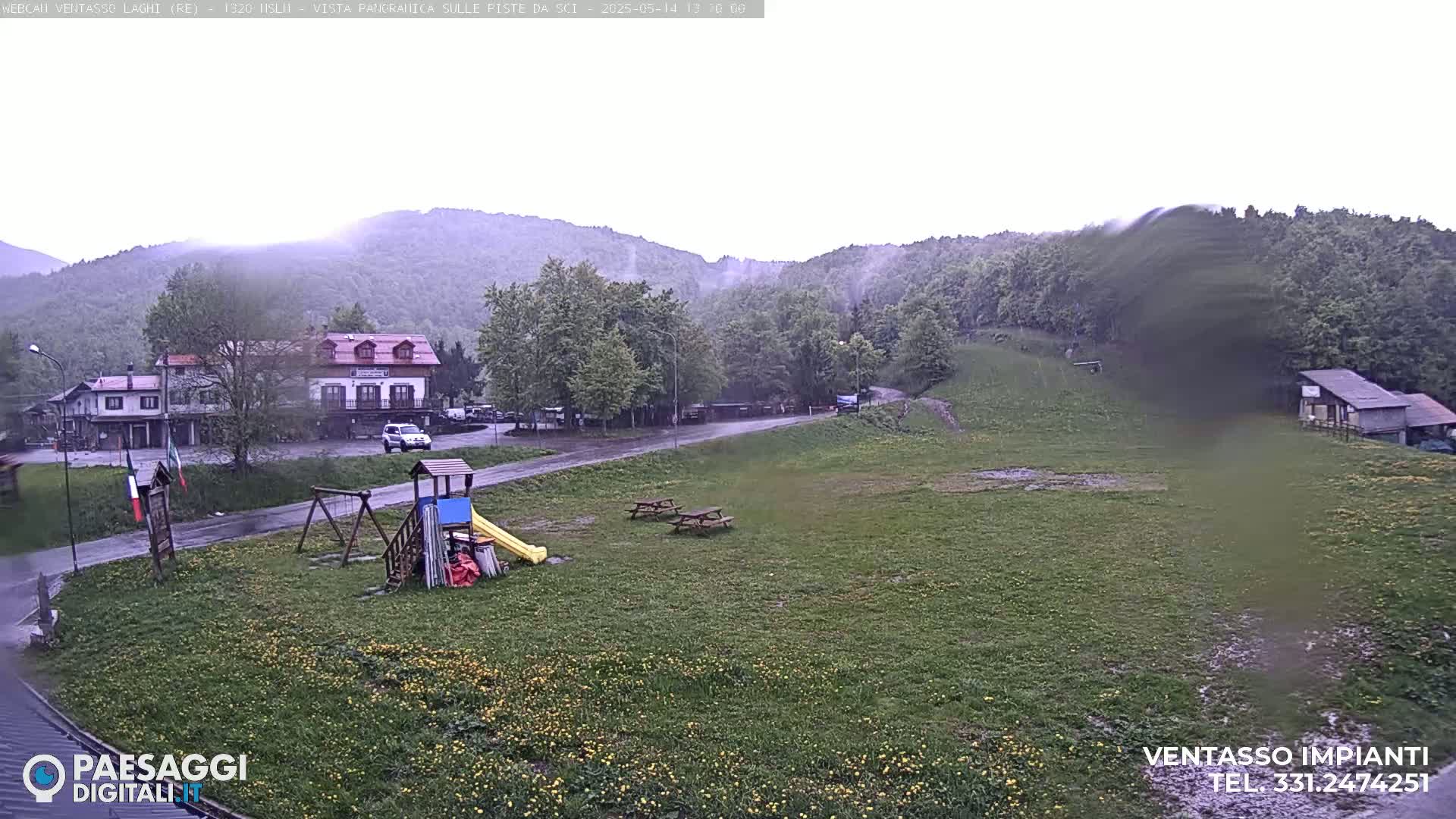 A rain-soaked playground and grassy field sit before a village nestled in a mountain valley under an overcast sky.