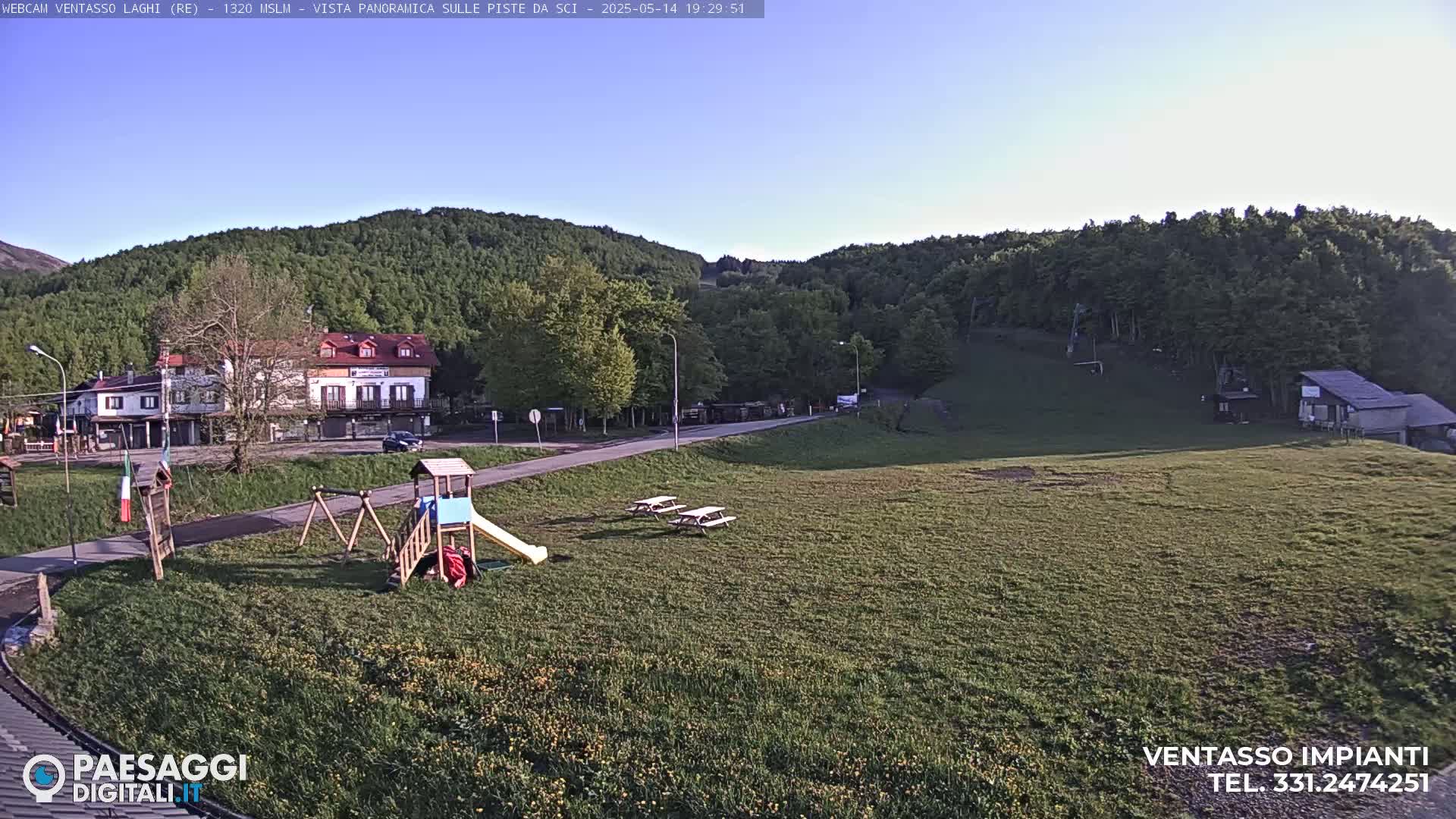 A grassy field with a playground, picnic tables, and a few buildings sits below a hillside covered in lush green trees under a clear blue sky.