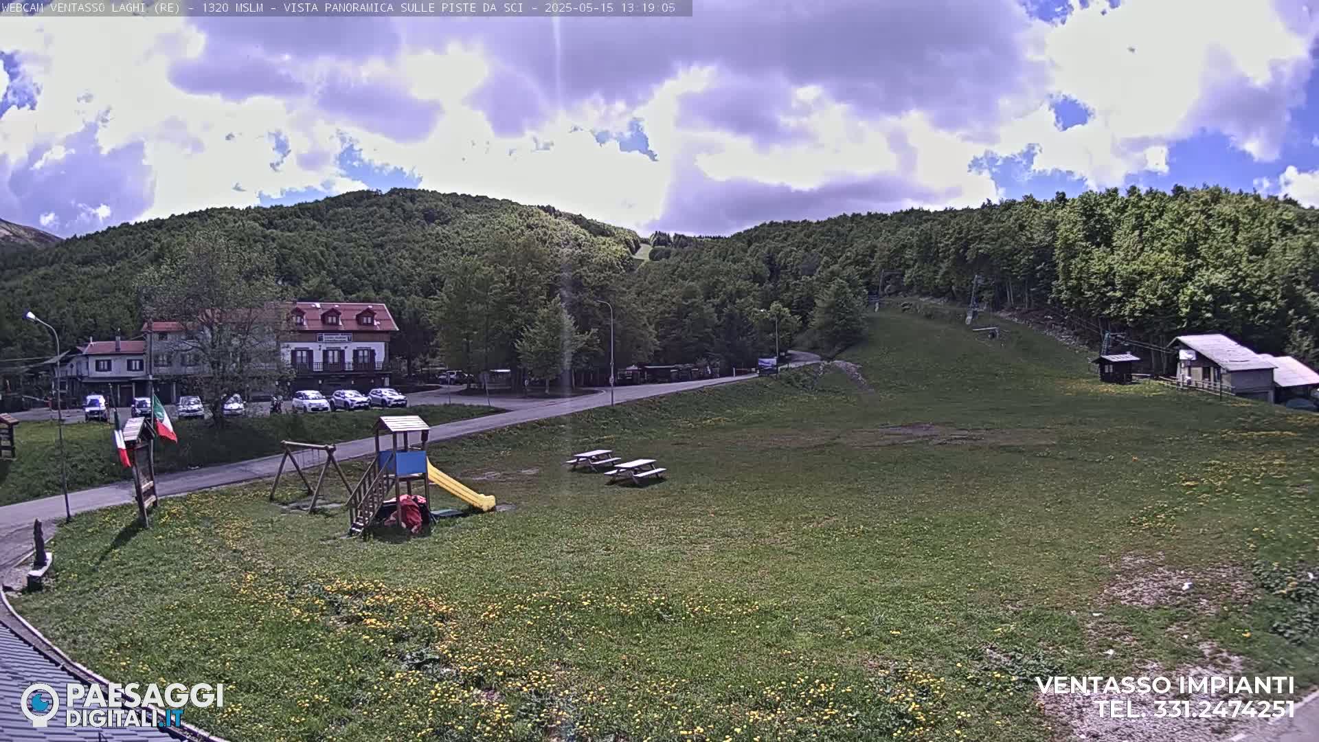 A partly cloudy day reveals a mountainside scene with a grassy area featuring a playground and picnic tables near a building and parking lot, all bordered by a forest.
