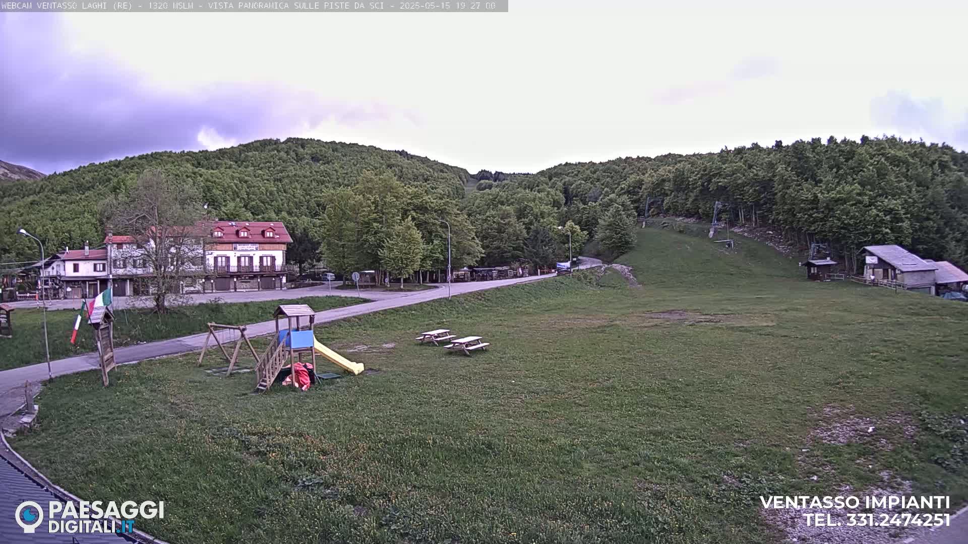 A playground and picnic tables sit on a grassy area near a building and ski lift against a backdrop of green hills under an overcast sky.