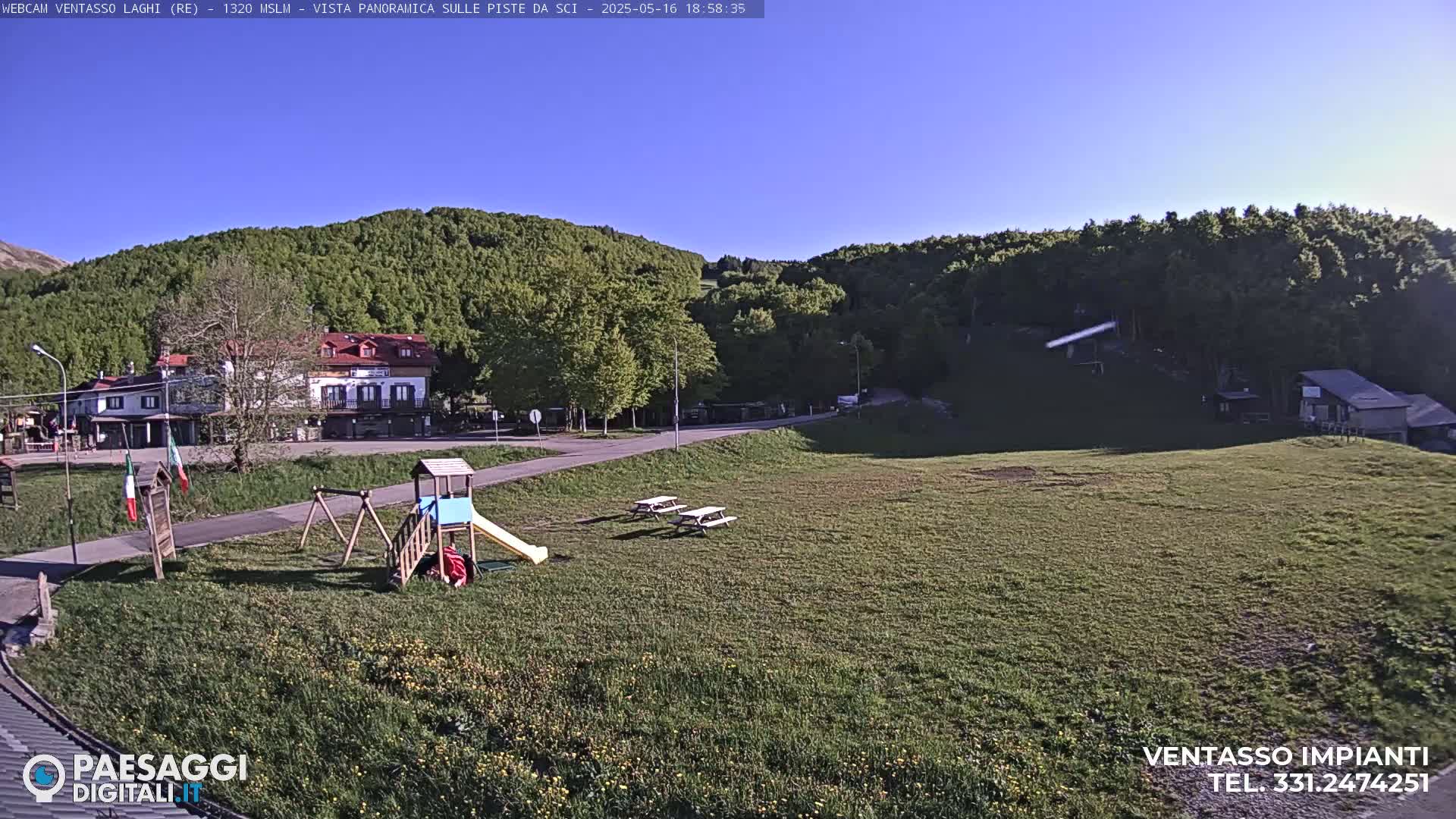 A grassy field with a playground, picnic tables, and a building is situated below a hillside covered in lush green trees under a clear blue sky.