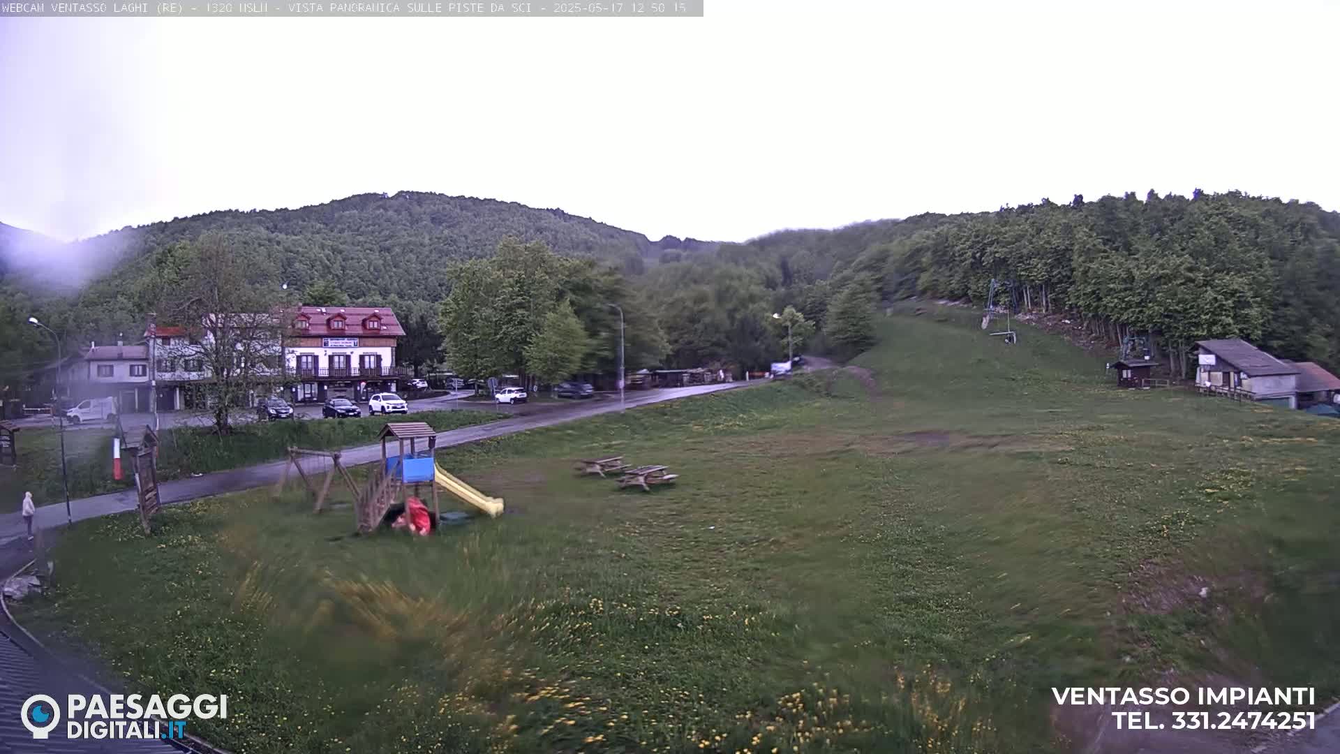 A rainy day shows a playground, picnic tables, a small building and a larger hotel-like building, situated in a grassy area near a forested hillside.