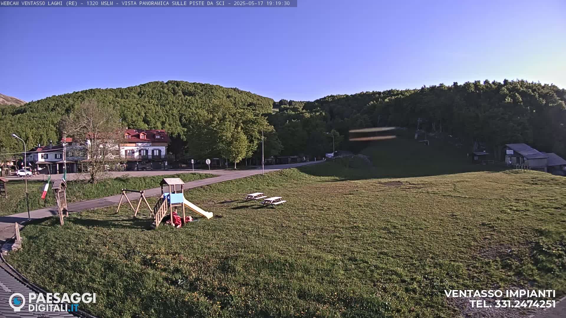 A playground and picnic tables sit on a grassy area overlooking a small village nestled among green hills under a clear blue sky.