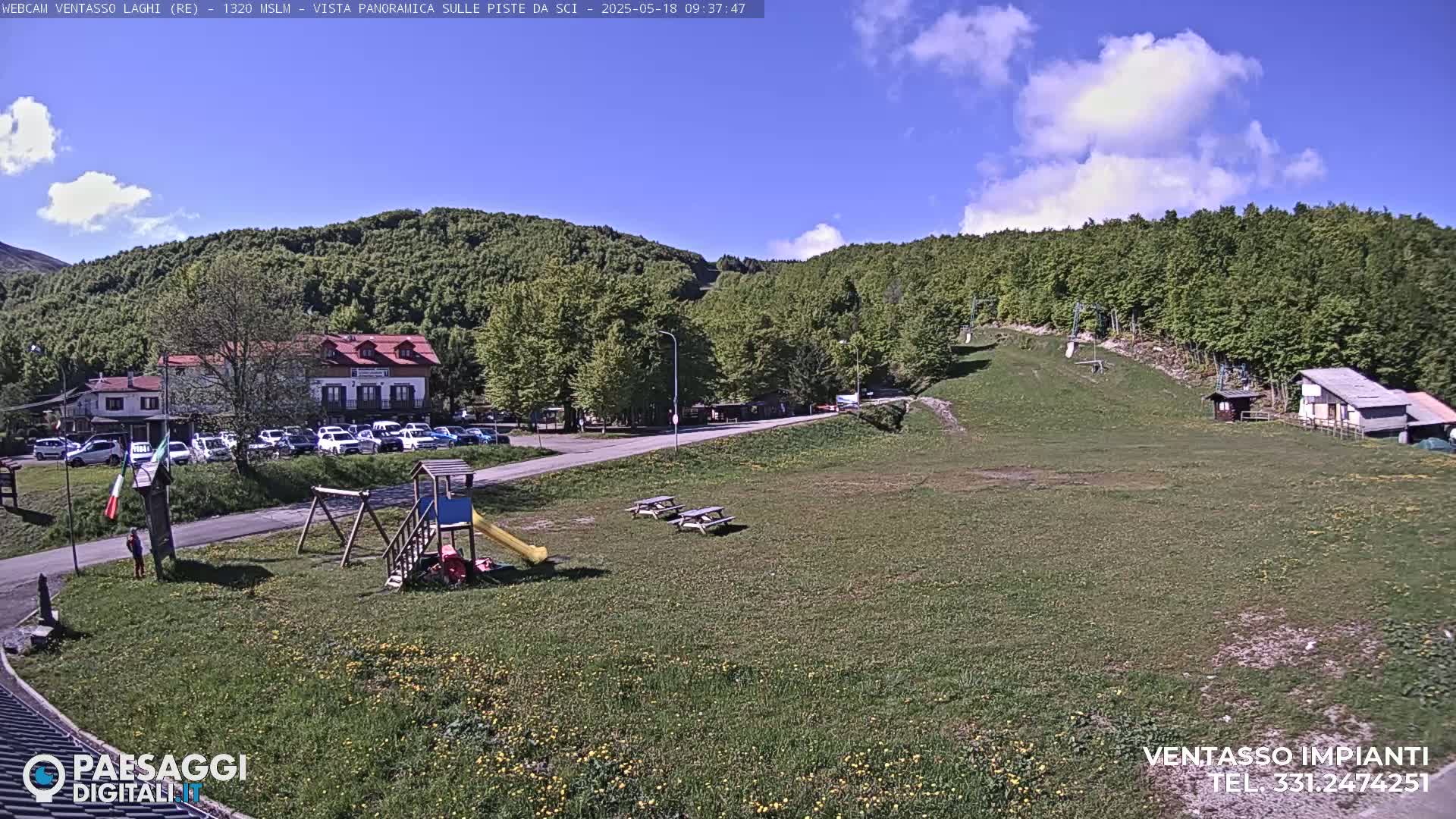A sunny day reveals a grassy area with a playground, picnic tables, a parking lot full of cars, a building, and a ski slope nestled among a lush green forest on a hillside.