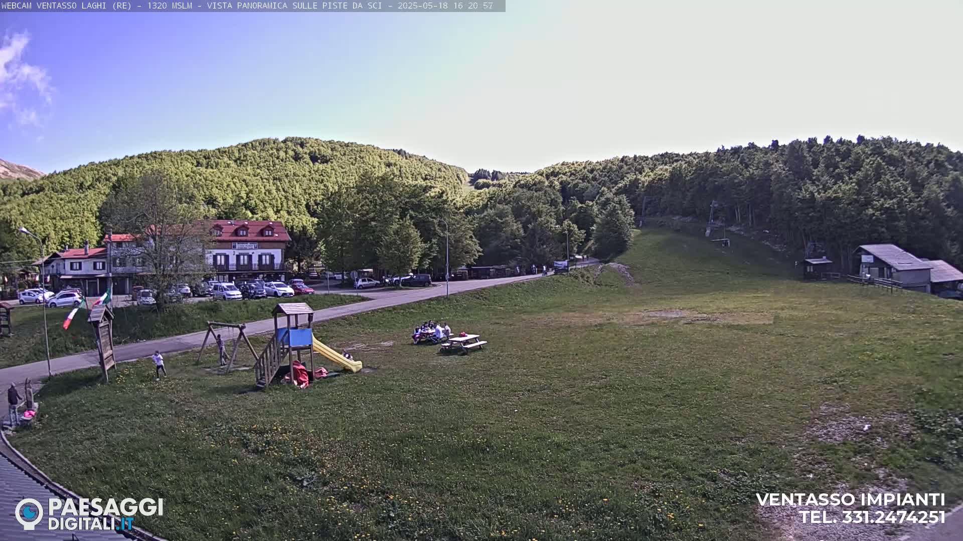 A sunny day reveals a grassy area with a playground, picnic tables, a building, and a parking lot, all nestled within a verdant hillside.