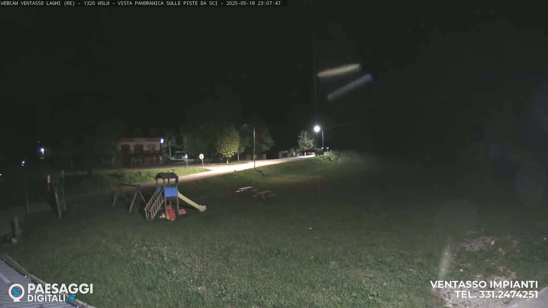 A nighttime view of a playground and picnic tables in a grassy area, illuminated by streetlights under a dark sky.