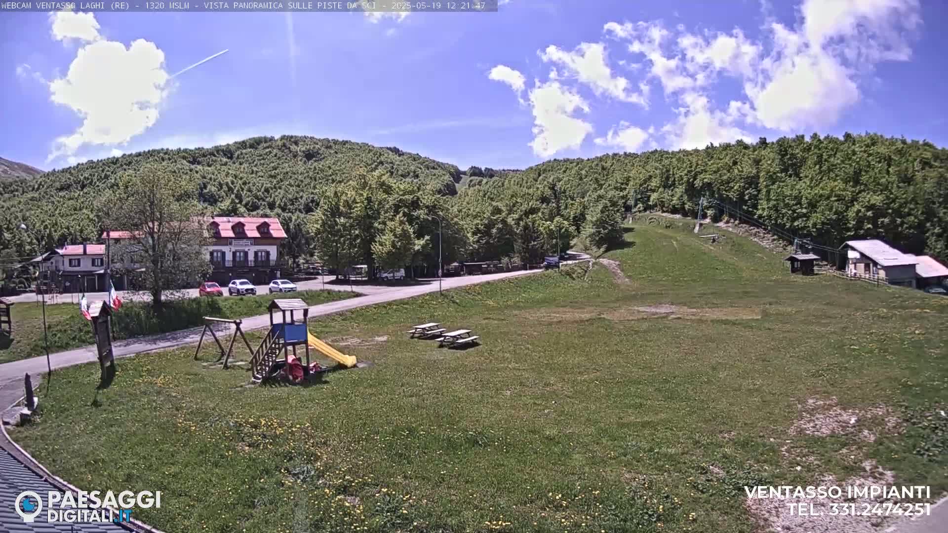 A mostly sunny day reveals a grassy area with a playground, picnic tables, a small building, and a parking lot near a forested hillside.