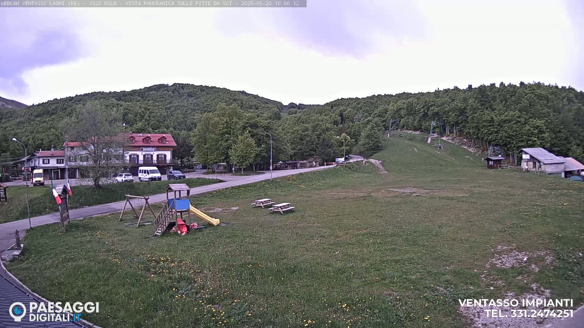 A playground and picnic tables sit in a grassy area near buildings and a ski lift against a backdrop of a lush green hill under an overcast sky.