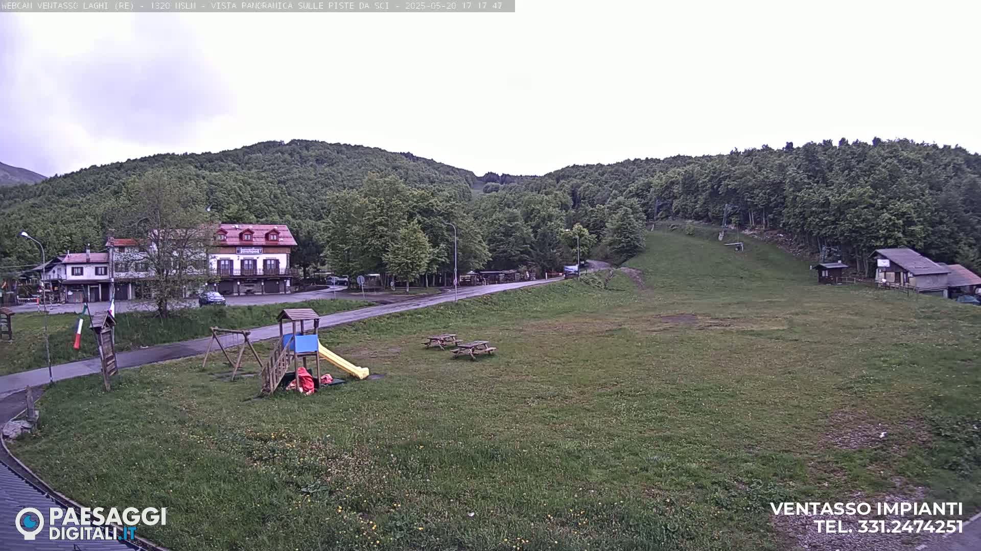 A playground and picnic tables sit on a grassy field in front of buildings and a forested hillside under an overcast sky.