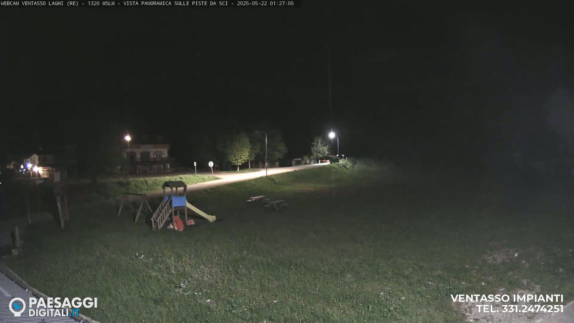 A nighttime view of an empty playground near a road, lit by streetlights, with a grassy area extending into the darkness.