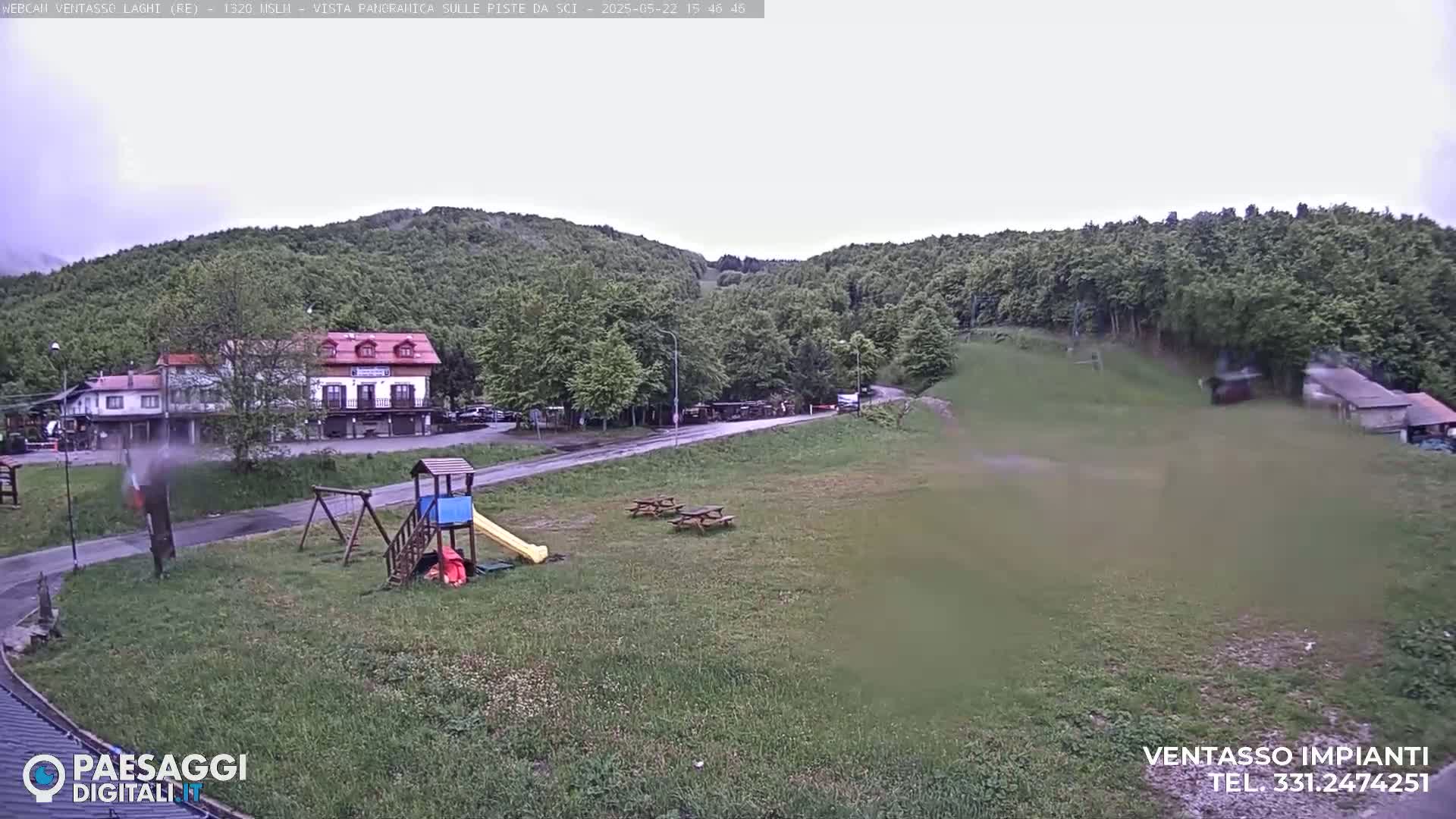 A playground with a slide and swings sits near a building in a grassy area, with overcast skies and a wooded hillside in the background.