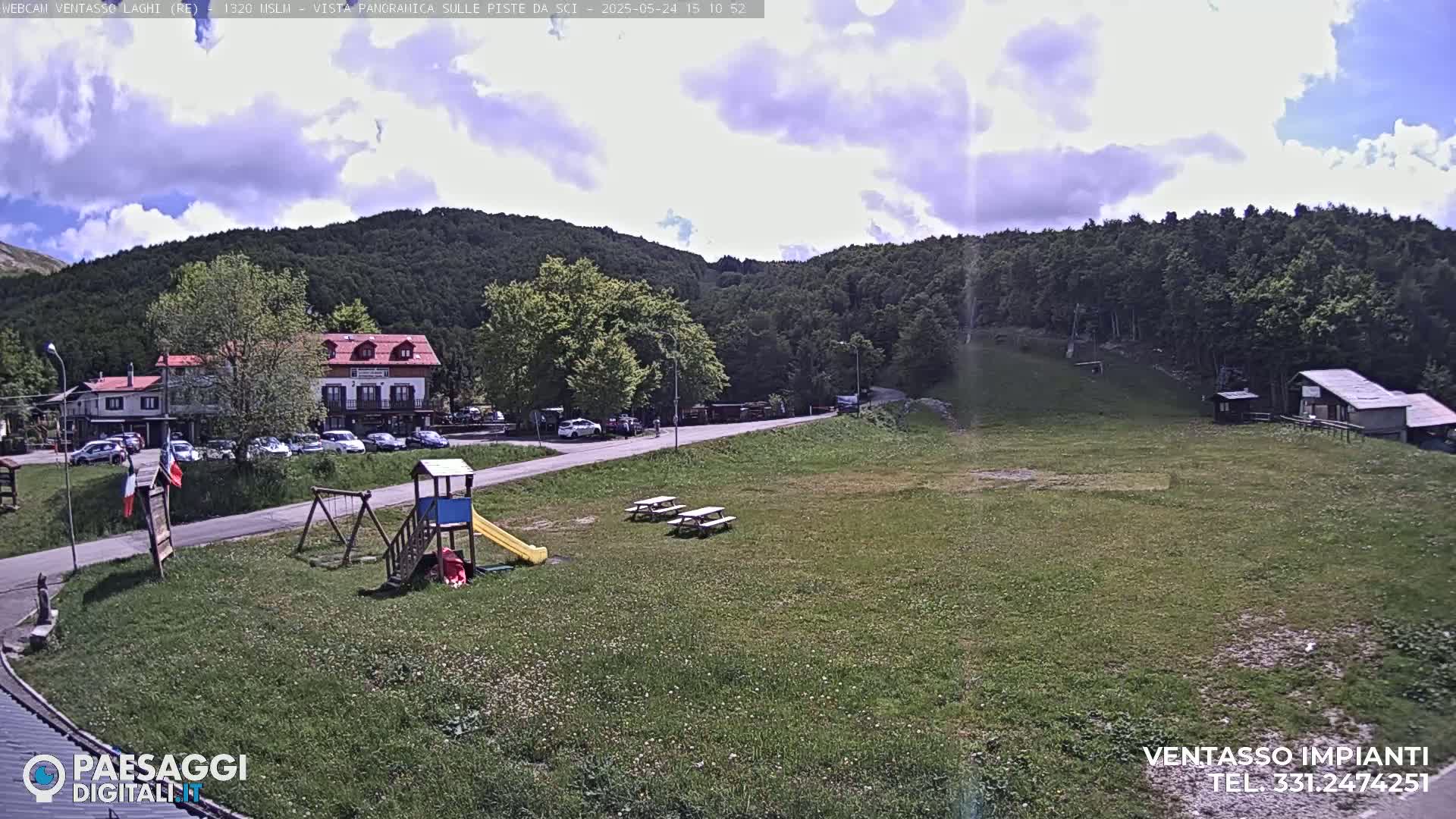 A partly cloudy day reveals a grassy area with picnic tables, a playground, and buildings nestled among a green, tree-covered mountain.