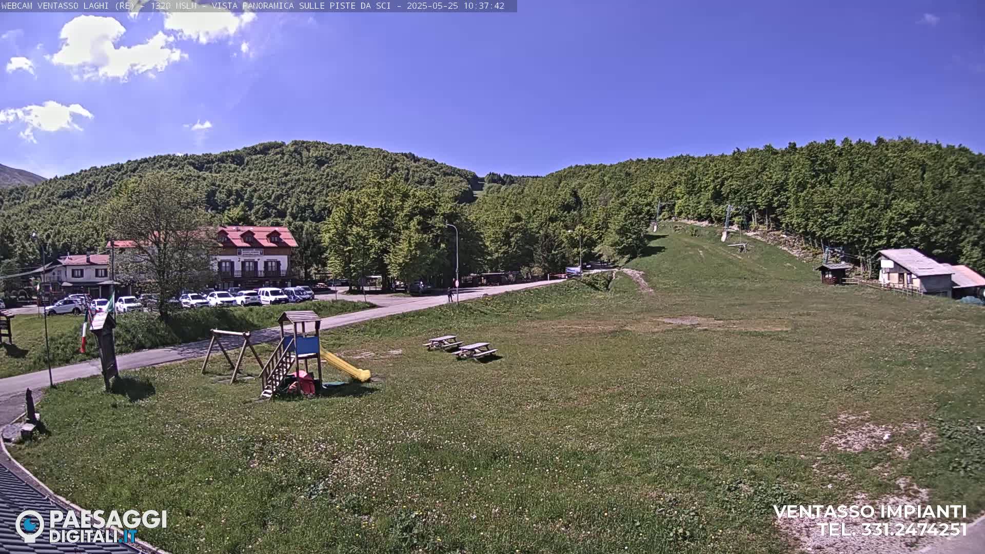 A sunny day at a mountain resort, showing a grassy area with picnic tables, a playground, buildings, and a ski lift in the background.