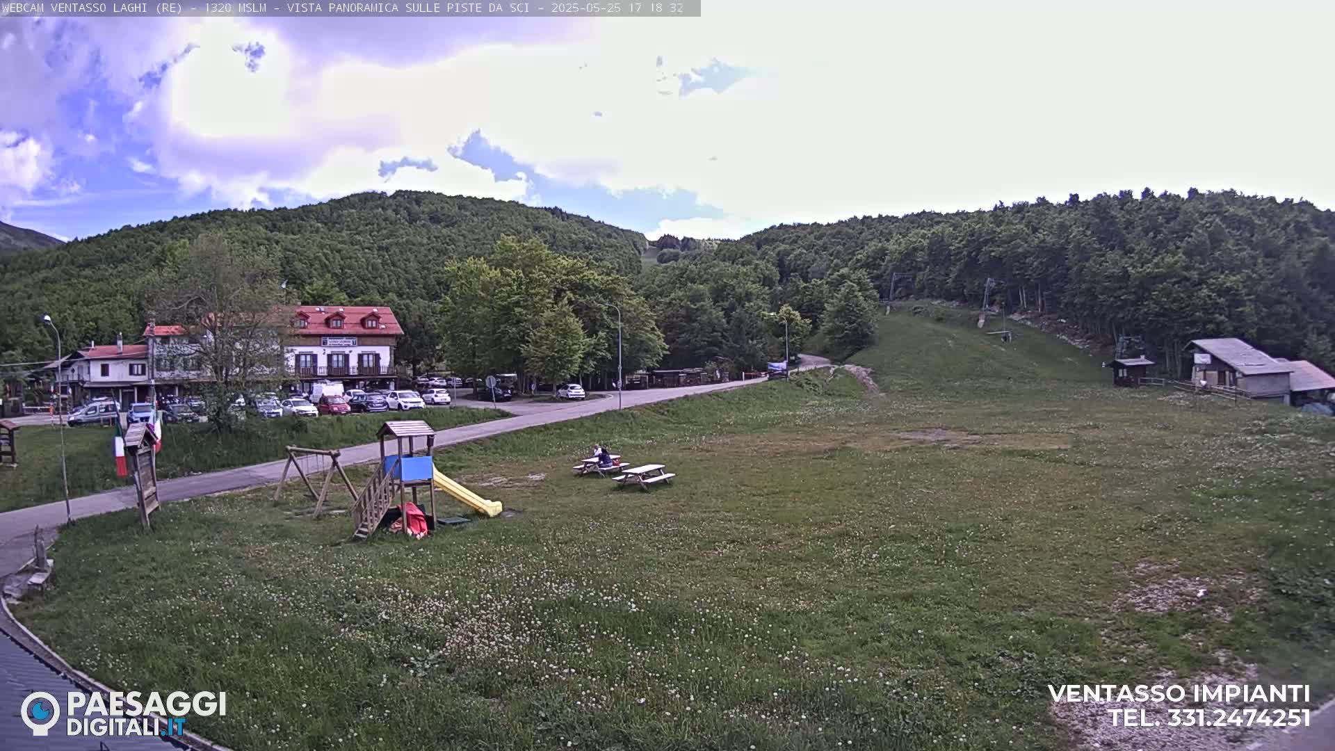 A partly cloudy day reveals a mountainside scene with a building, parking lot, playground, picnic tables, and grassy areas, all overlooking a ski slope.