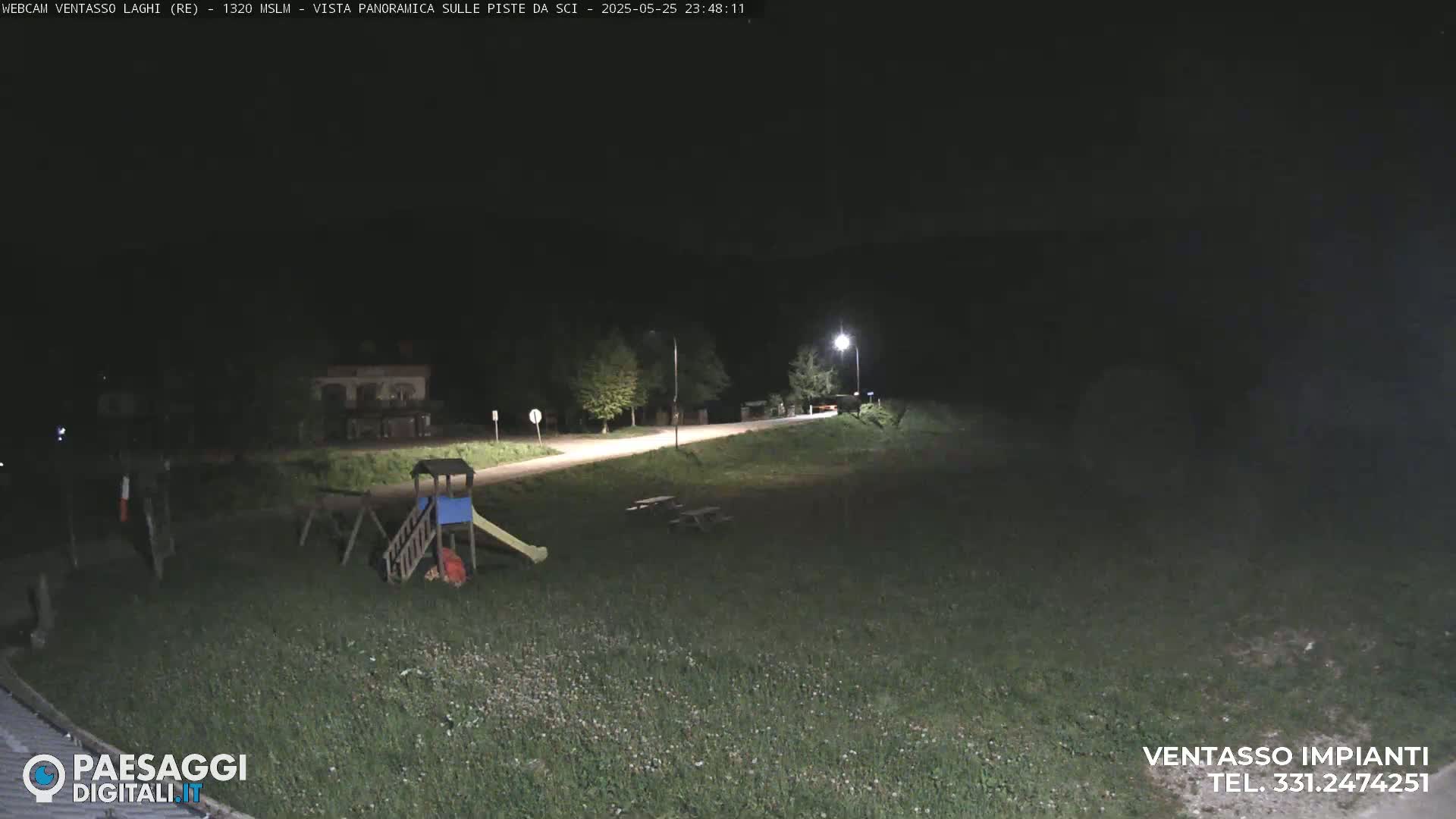 A nighttime view of a grassy area with a playground, picnic tables, and a road illuminated by streetlights.