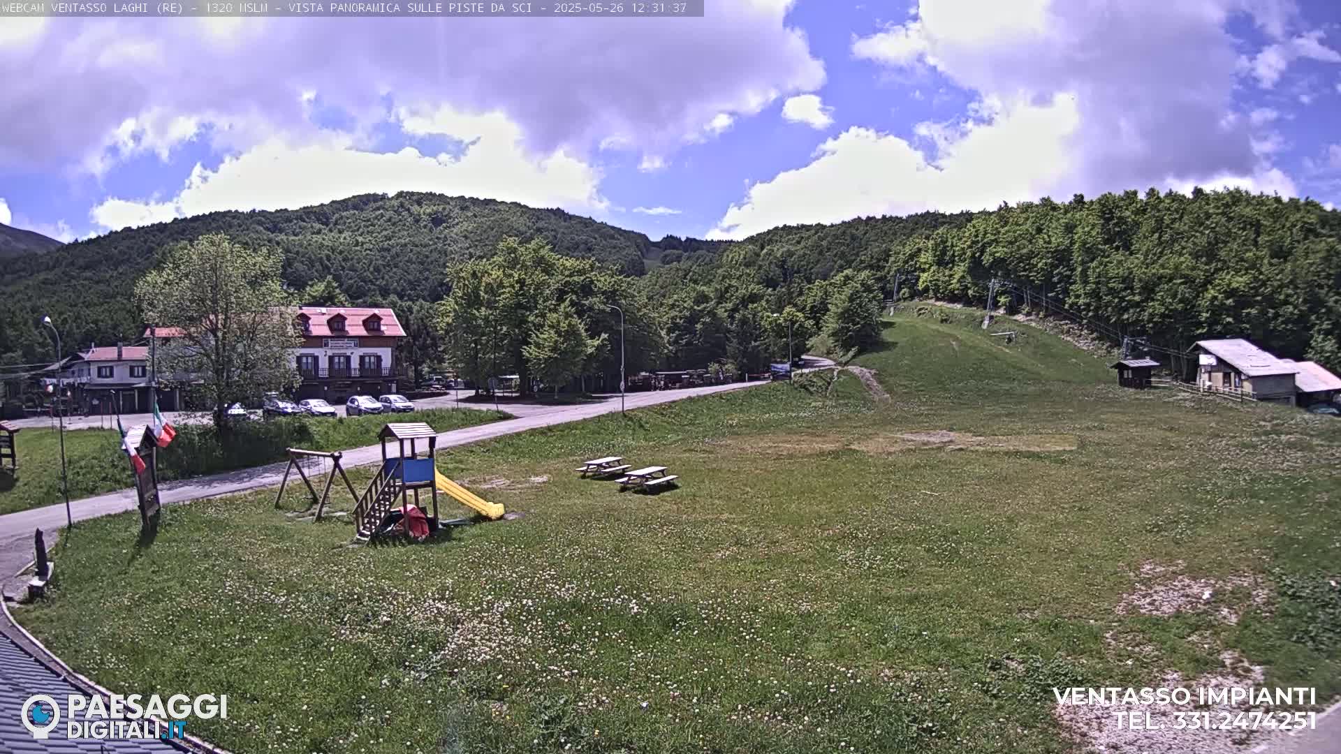 A sunny day reveals a grassy field with a playground, picnic tables, a few buildings, and a ski lift in the background, all nestled amidst green hills under a partly cloudy sky.