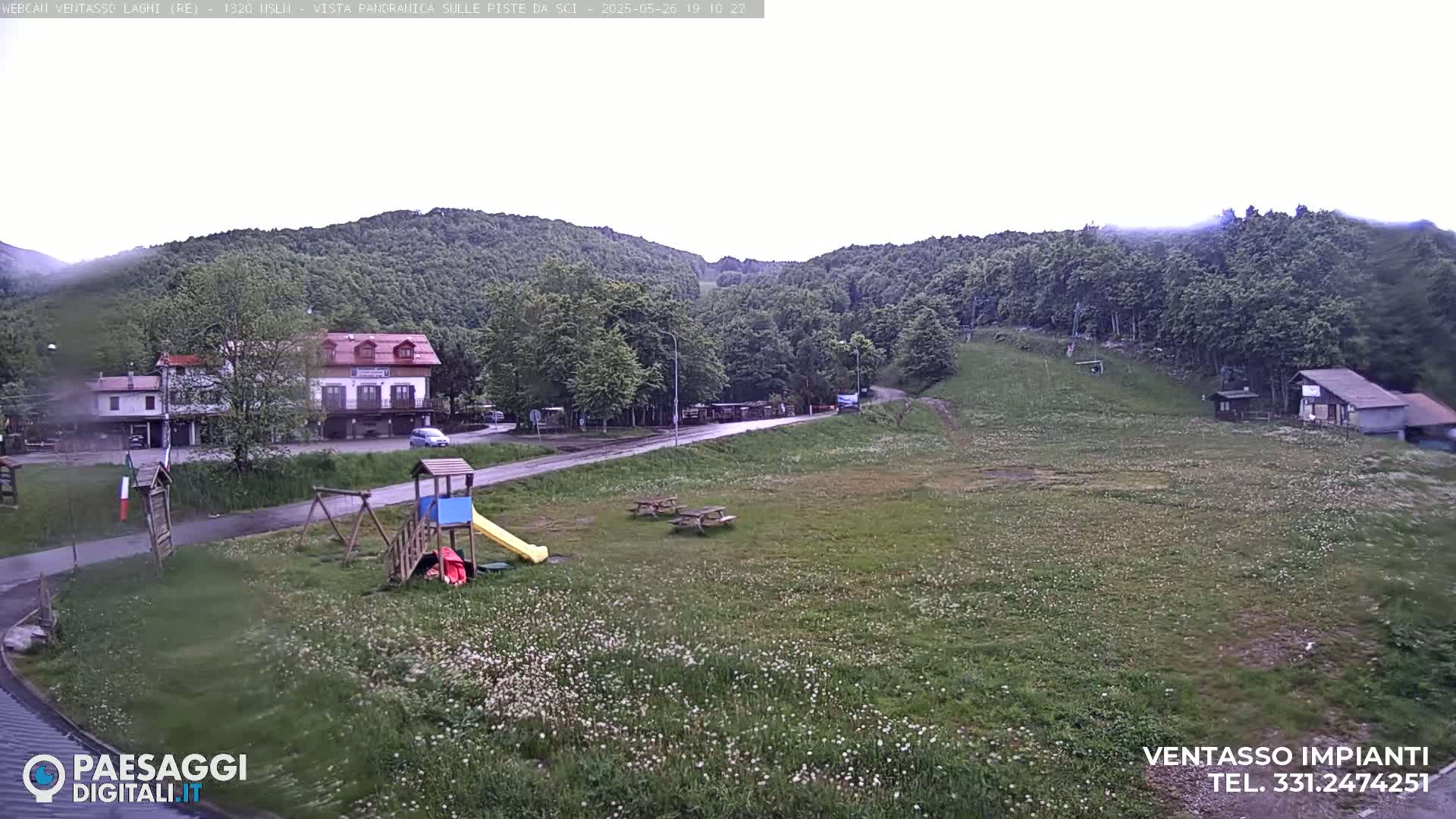 A wet, grassy area with a playground, picnic tables, and buildings nestled amongst green hills under an overcast sky.