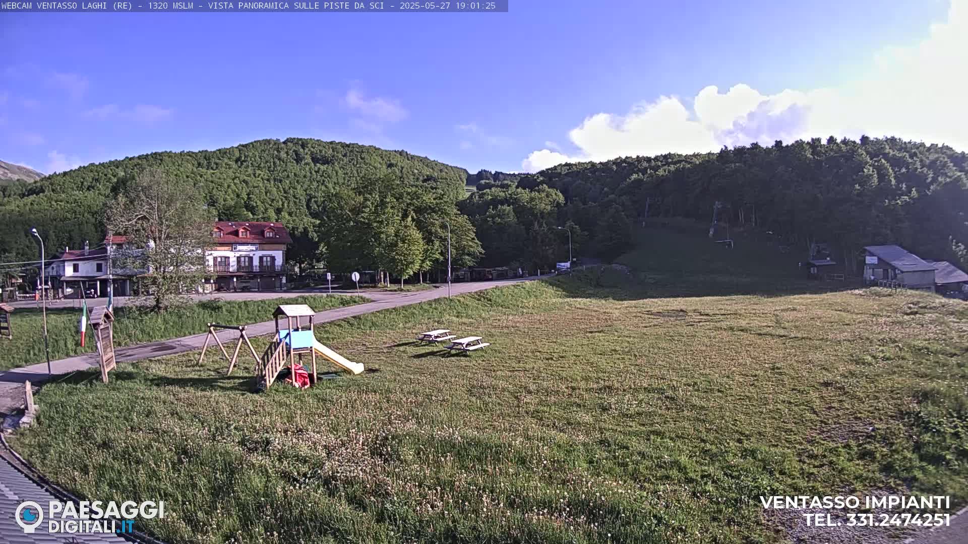 A sunny day reveals a grassy field with a playground, picnic tables, a few buildings, and a forest-covered mountain in the background.
