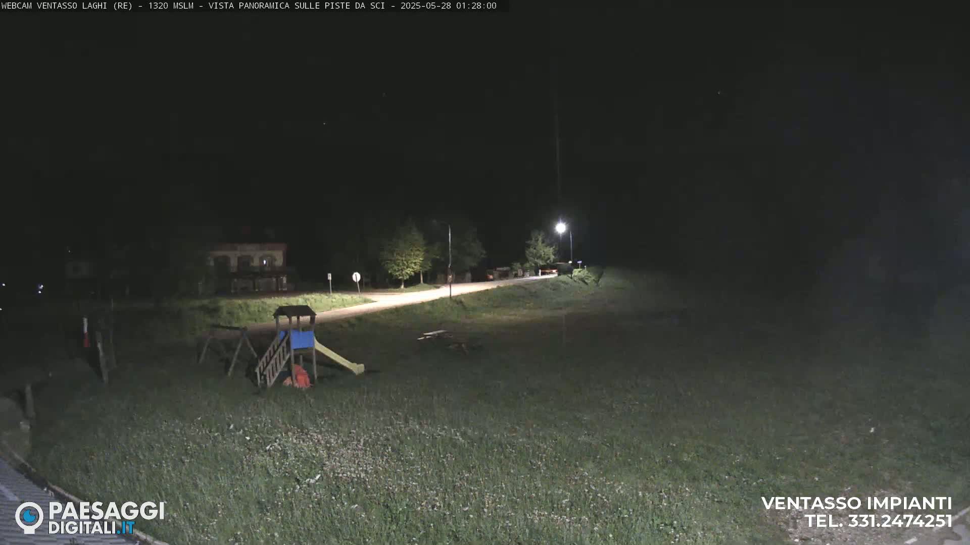 A nighttime view of a playground and a road next to a grassy field, illuminated by a streetlight.