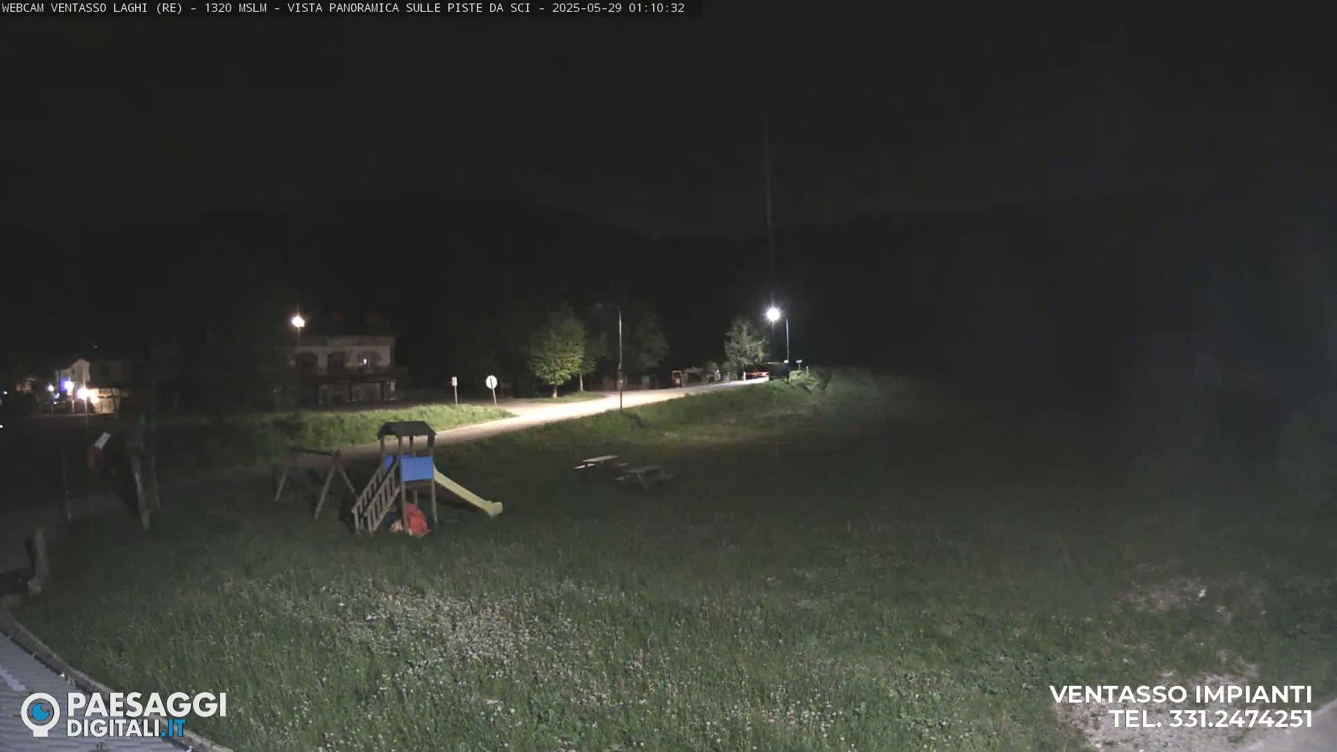 A nighttime view of a playground near a road, illuminated by streetlights, with grassy areas surrounding it.