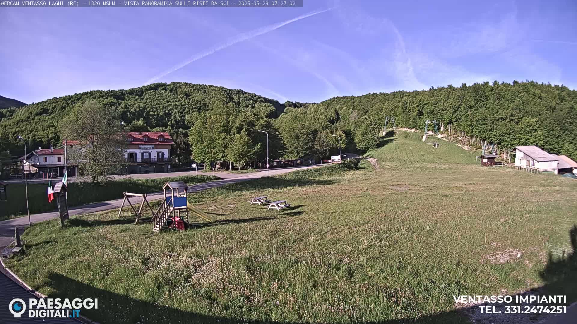 A sunny, clear day overlooks a grassy field with a playground, picnic tables, buildings, and a ski lift in the background, all surrounded by a lush green forest.