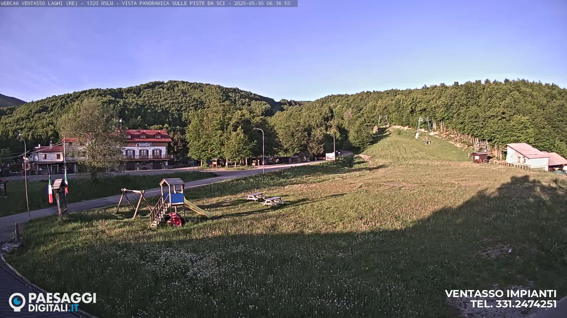A sunny view of a grassy field with a playground, buildings, and a forested hillside in the background.