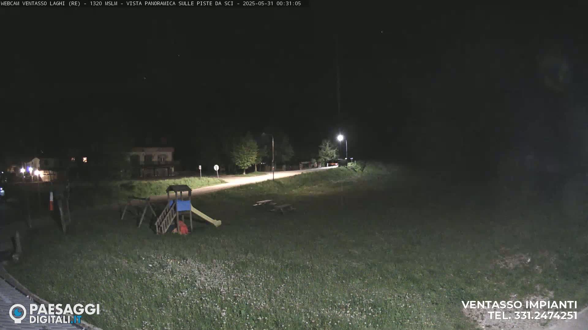A nighttime view of a grassy area with a playground, picnic tables, and a road leading to some buildings under a dark, clear sky.