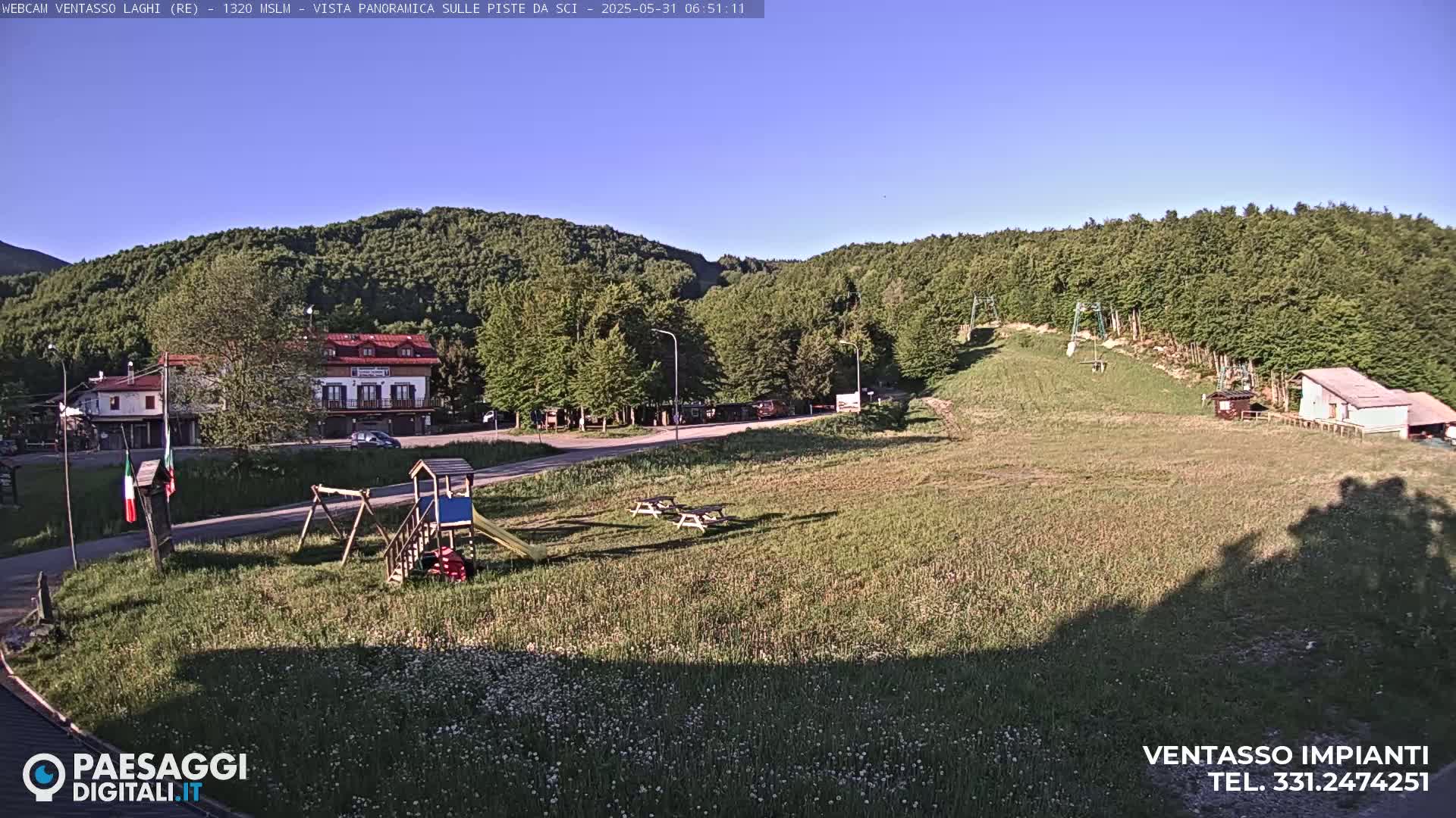 A grassy field with a playground, picnic tables, and a building sits below a hill covered in lush green trees under a clear blue sky.