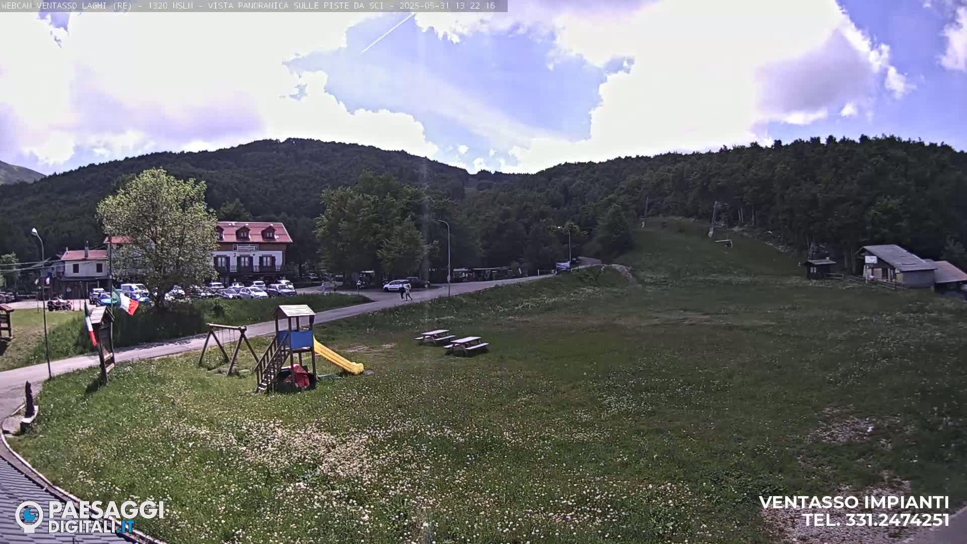 A partly sunny day reveals a grassy area with a playground, picnic tables, a parking lot with cars, a building, and a forested hillside in the background.